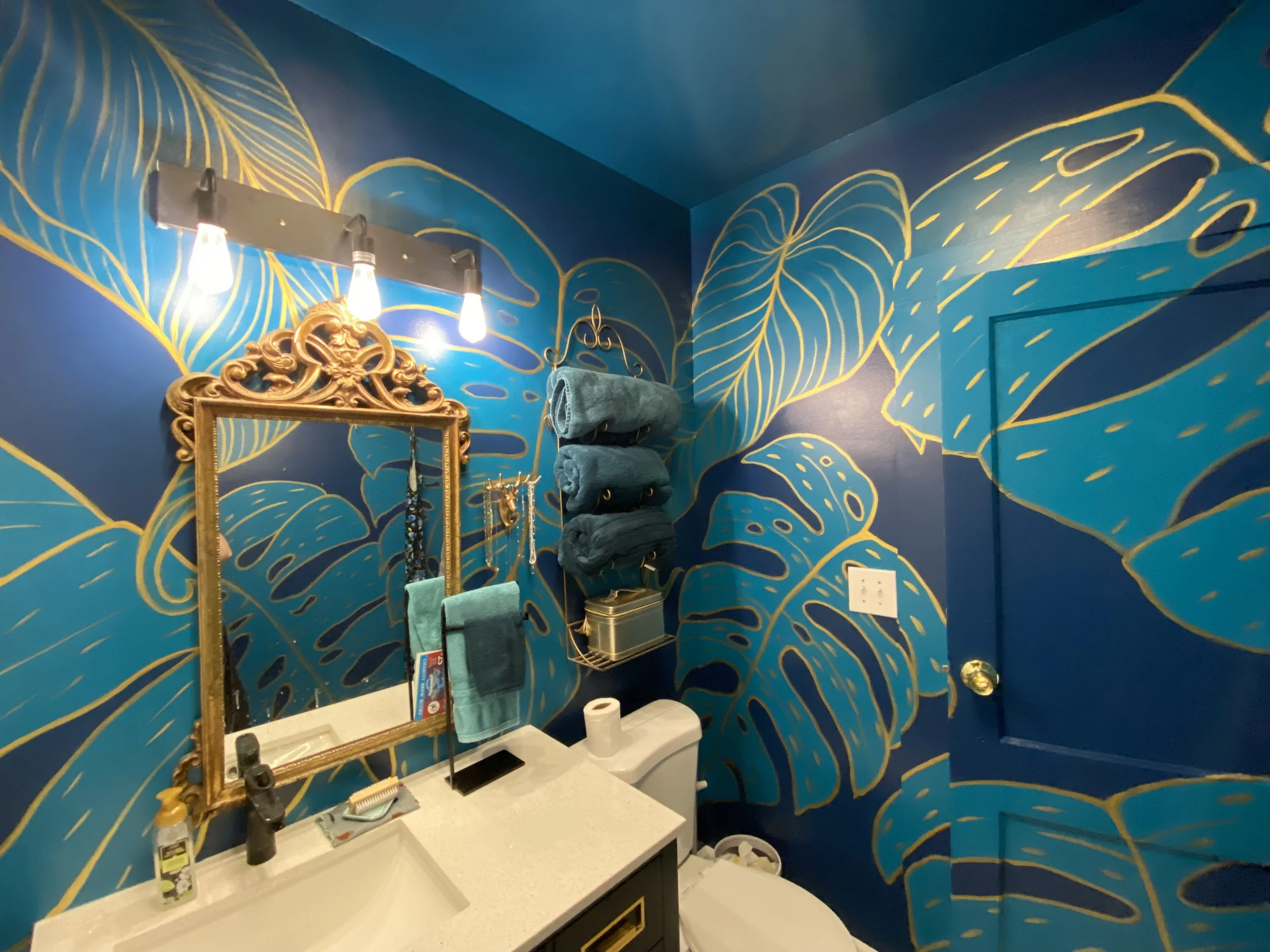 Bathroom with vibrant blue walls decorated with large yellow leaf patterns, with a gold ornate mirror, three light bulbs above, and a towel rack holding multiple teal towels above a white toilet and sink.