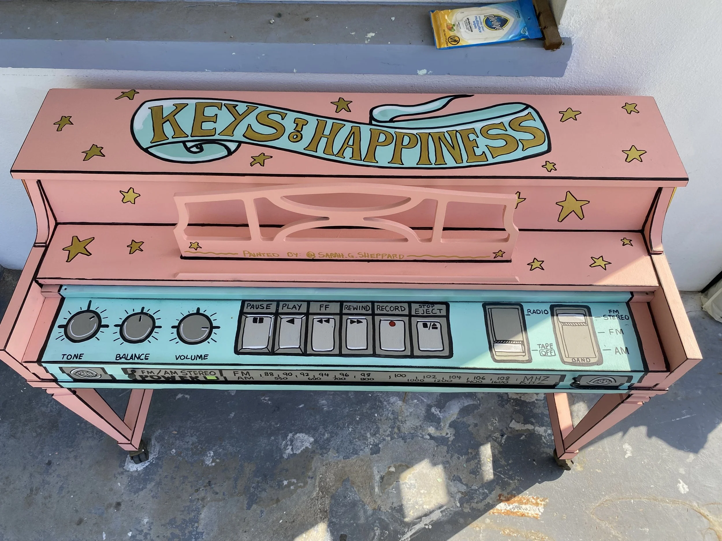 Pink painted vintage radio-shaped table with control dials labeled tone, balance, and volume, and buttons labeled pause, play, fast forward, rewind, record, stop, eject, and others, decorated with yellow stars and a large banner reading 'KEYS TO HAPP