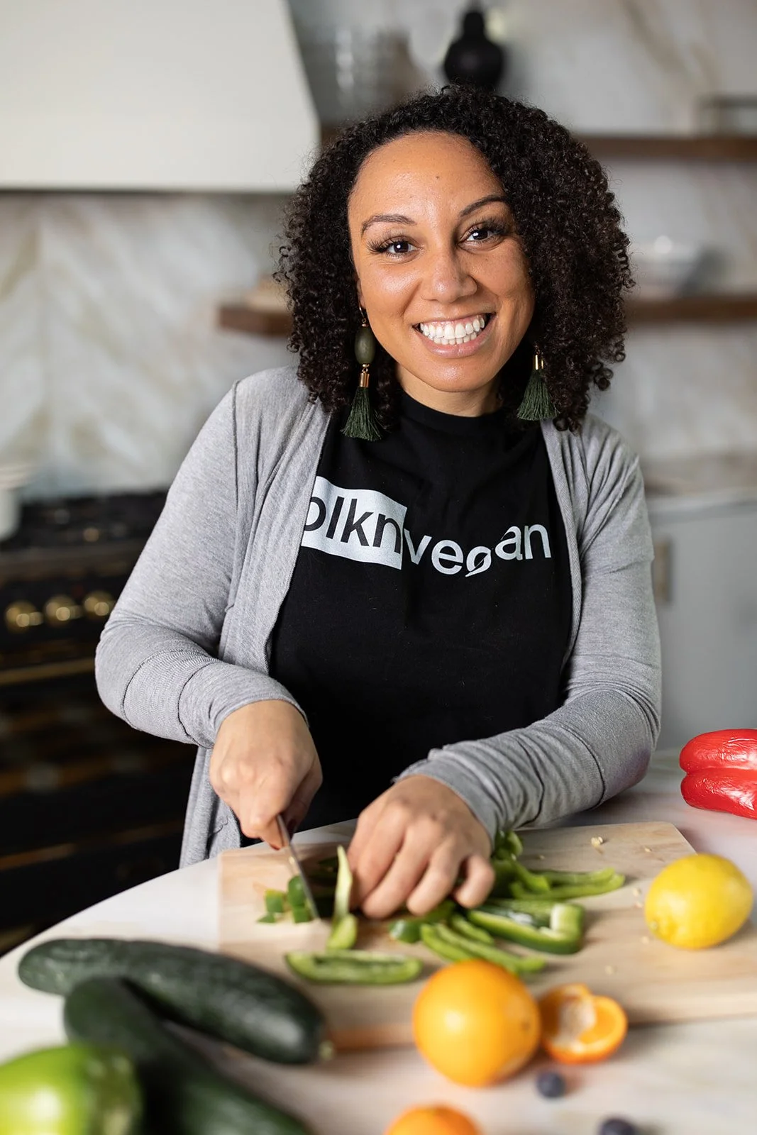 A woman with curly dark hair, smiling, chopping green vegetables on a cutting board in a kitchen. She is wearing a black t-shirt with the word 'vegan' and a light gray cardigan. Several cucumbers, oranges, a lemon, and a red bell pepper are on the counter.