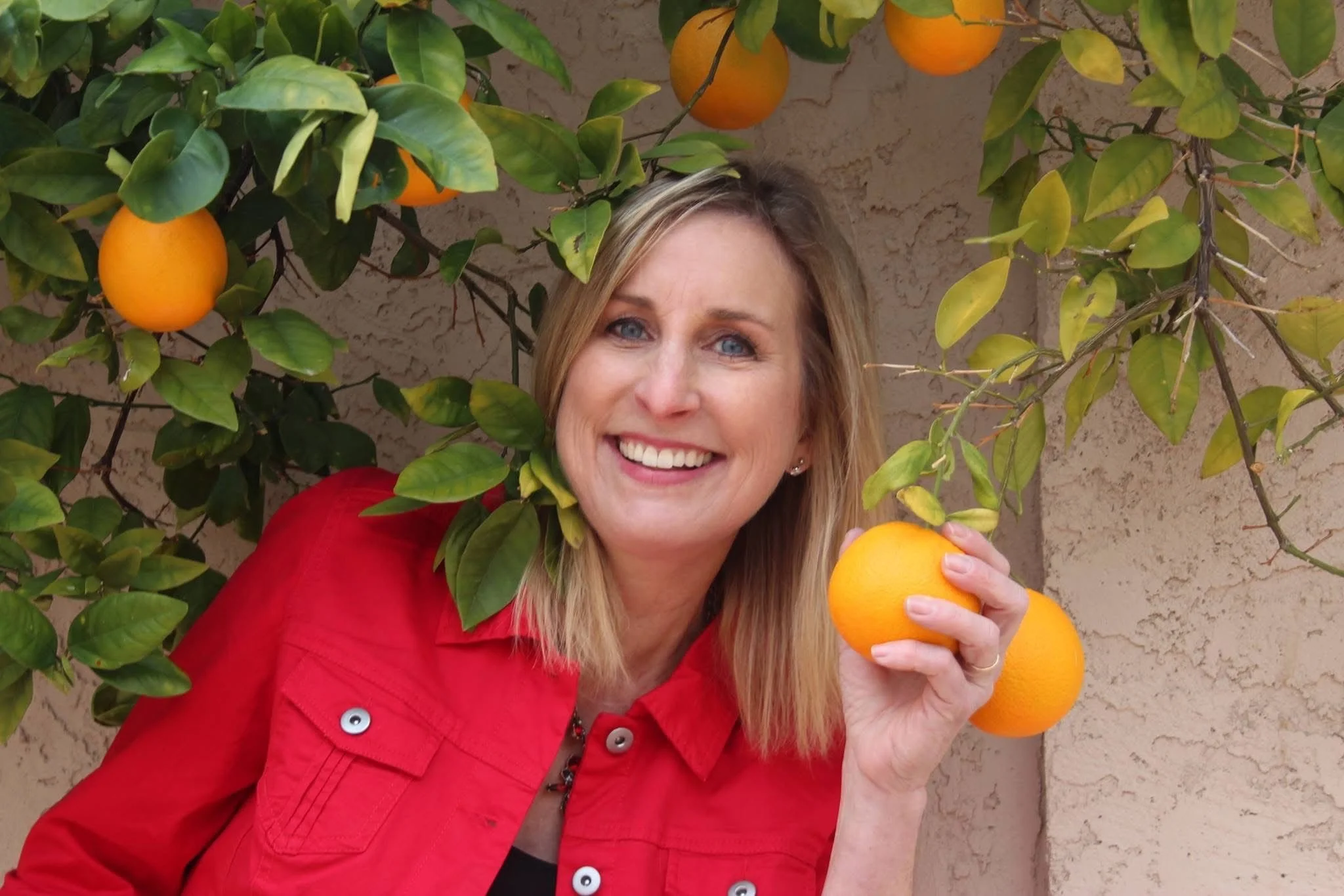 A woman smiling and holding an orange in front of an orange tree with ripe oranges and green leaves.