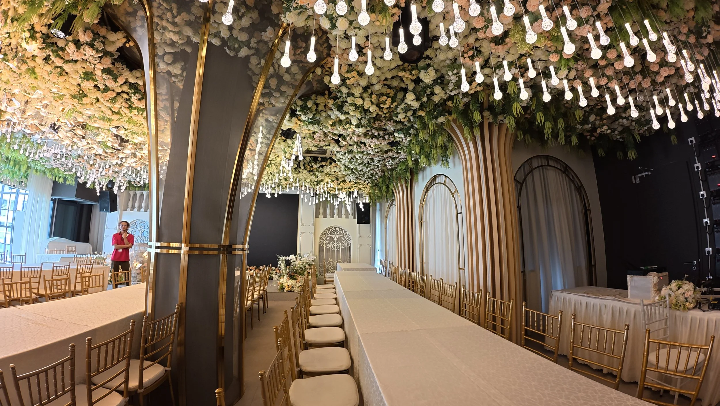 Elegant event hall decorated with hanging light bulbs, floral ceiling arrangement, and gold chairs around tables, with a person standing near a stage.