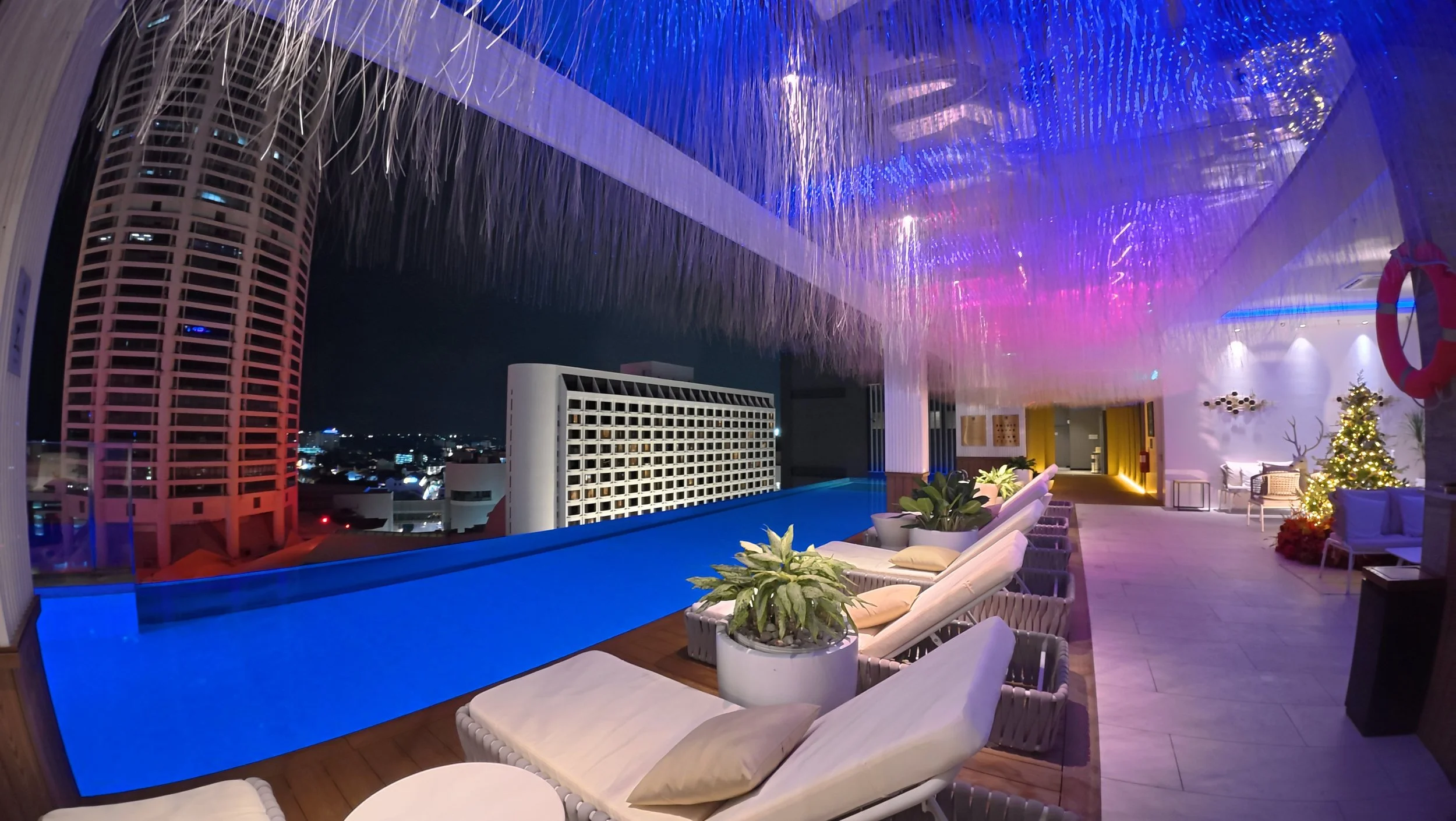 Nighttime rooftop pool area with lounge chairs, potted plants, and a decorated Christmas tree, under a thatched roof with colorful lights