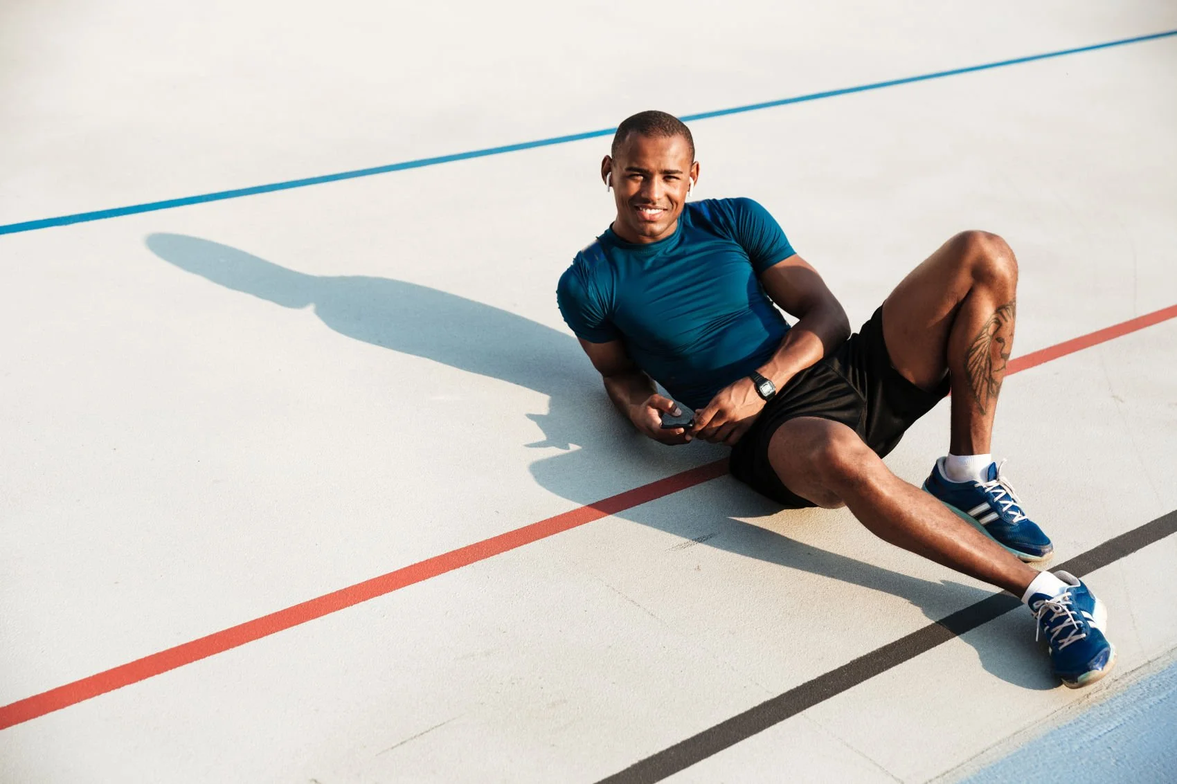 Man sitting on a track with earphones, wearing a blue shirt, black shorts, and blue sneakers.