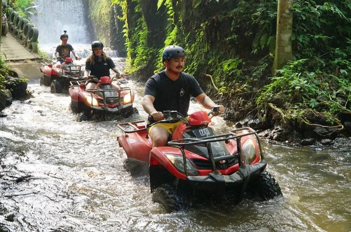 Three people riding red ATVs through a shallow stream surrounded by lush greenery, with a waterfall in the background.