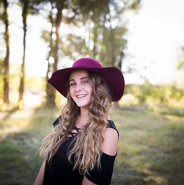 Woman wearing a burgundy hat and black dress, smiling in a sunny outdoor setting with trees in the background.