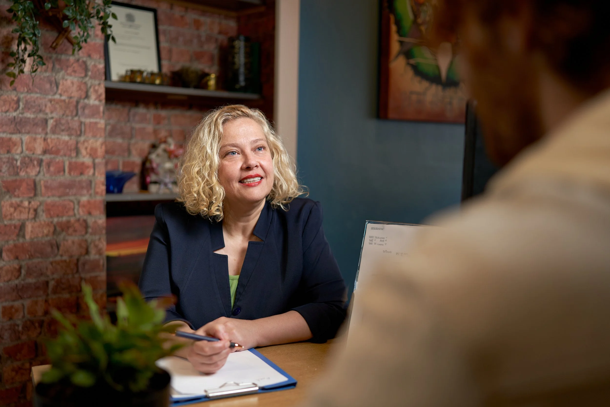A woman with curly blonde hair wearing a dark blazer sitting at a table and talking to a man, with a notebook in her hand and a brick wall in the background.