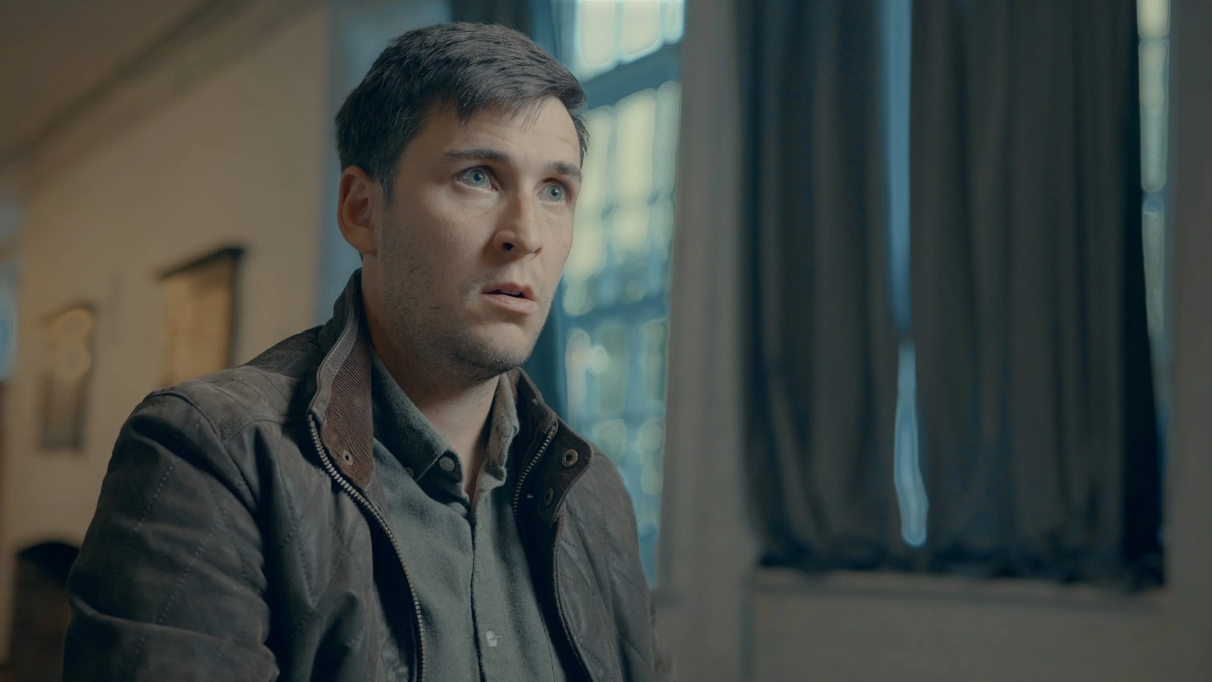 A man with dark hair and blue eyes looking confused or surprised, sitting indoors near a window with blue curtains.