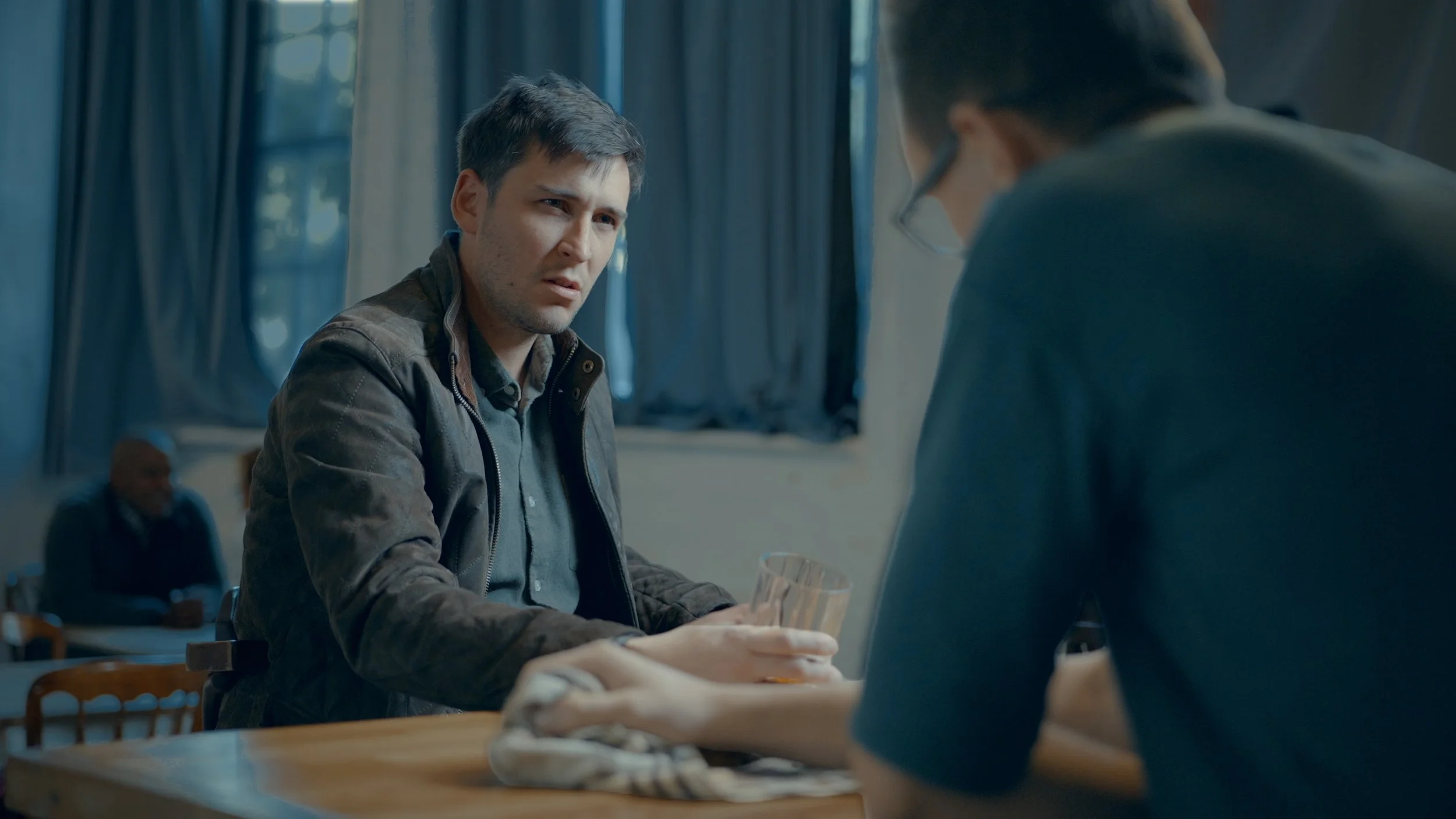 A young man with dark hair, wearing a gray shirt and brown jacket, looks confused or concerned while speaking to another person who has short dark hair, glasses, and is wearing a dark shirt. The scene appears to be indoors, with curtains and a window