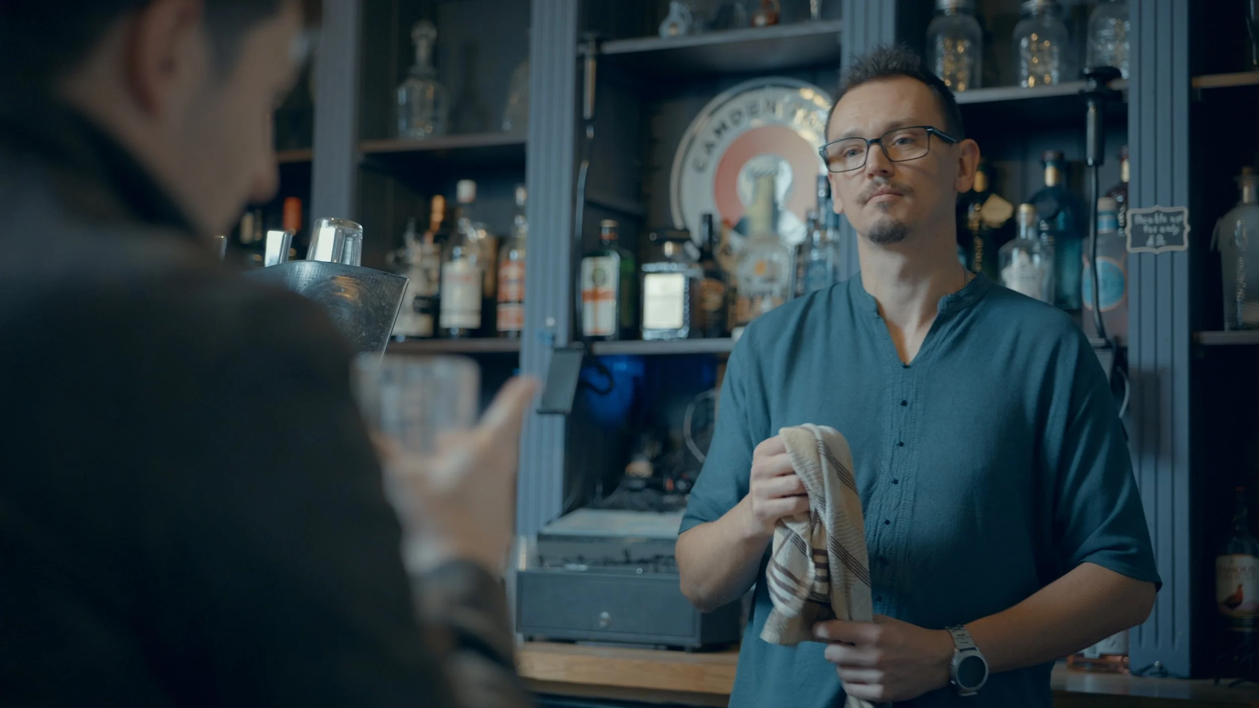 A man with glasses and a goatee, holding a striped cloth towel, stands behind a bar or counter, with shelves of bottles and bar equipment in the background. Another person is partially visible in the foreground, using a mobile device.