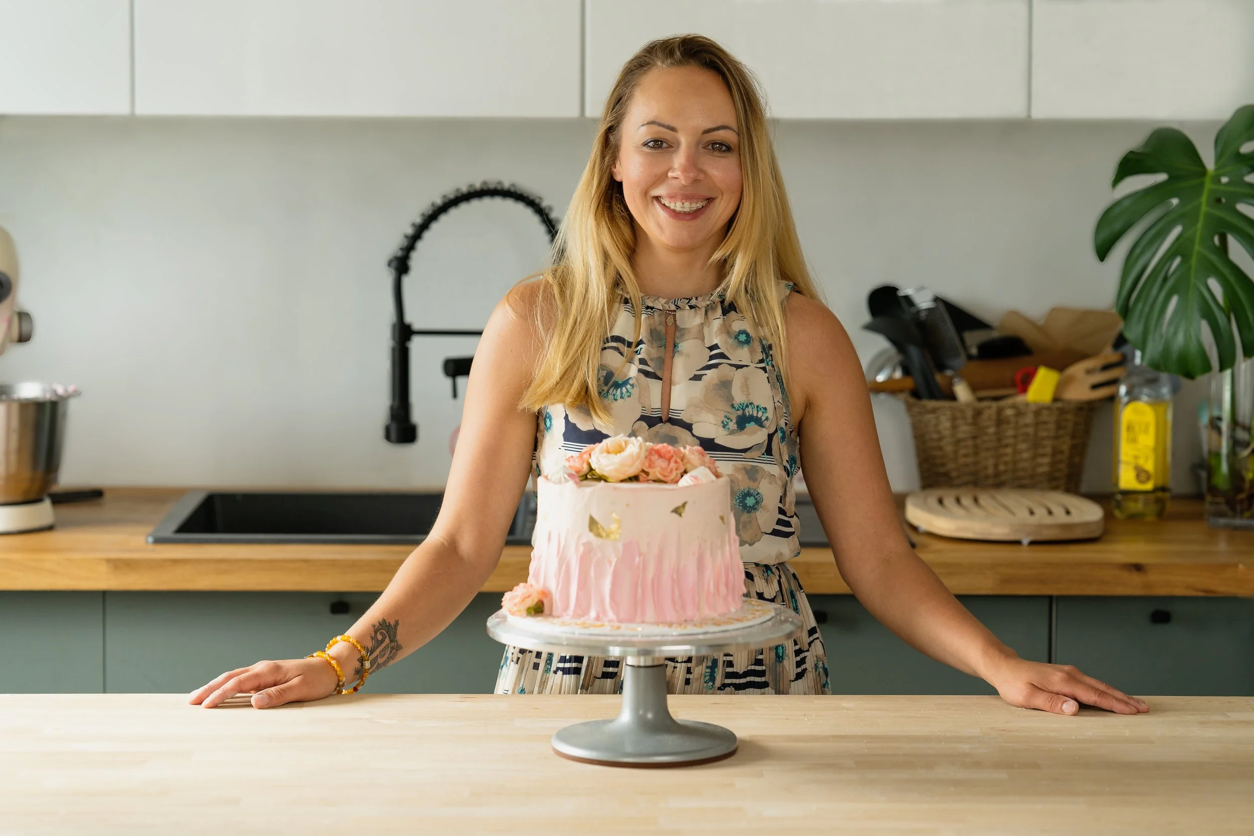 A woman with long blonde hair in a colorful dress smiling at the camera while standing behind a pink and white cake with flowers on top on a cake stand in a kitchen.