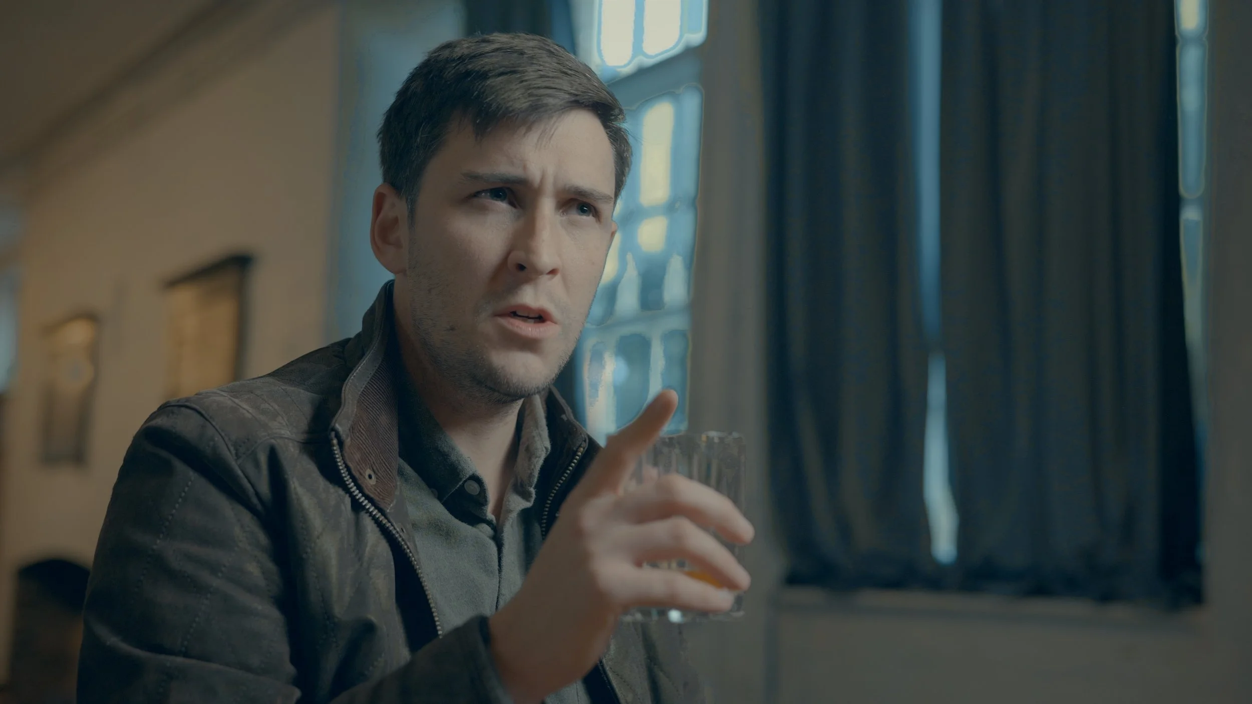 A man with short dark hair and a serious expression holding a glass of whiskey, sitting in a room with blue curtains and framed pictures on the wall.