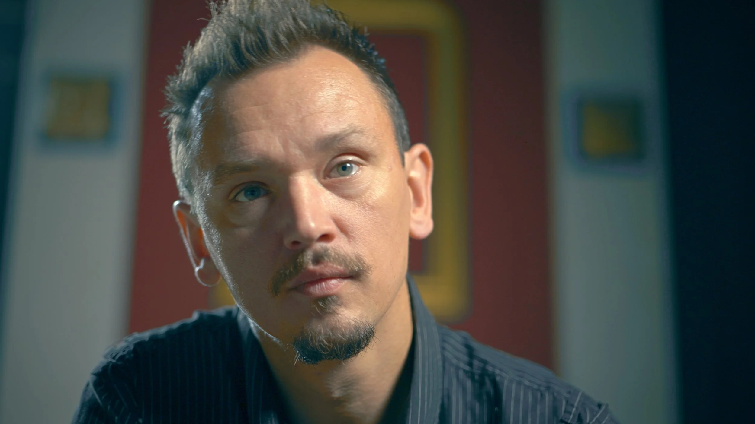 Close-up of a man with light skin, short dark hair, a beard, and blue eyes, wearing a dark striped shirt, inside a room with colorful artwork on the walls.