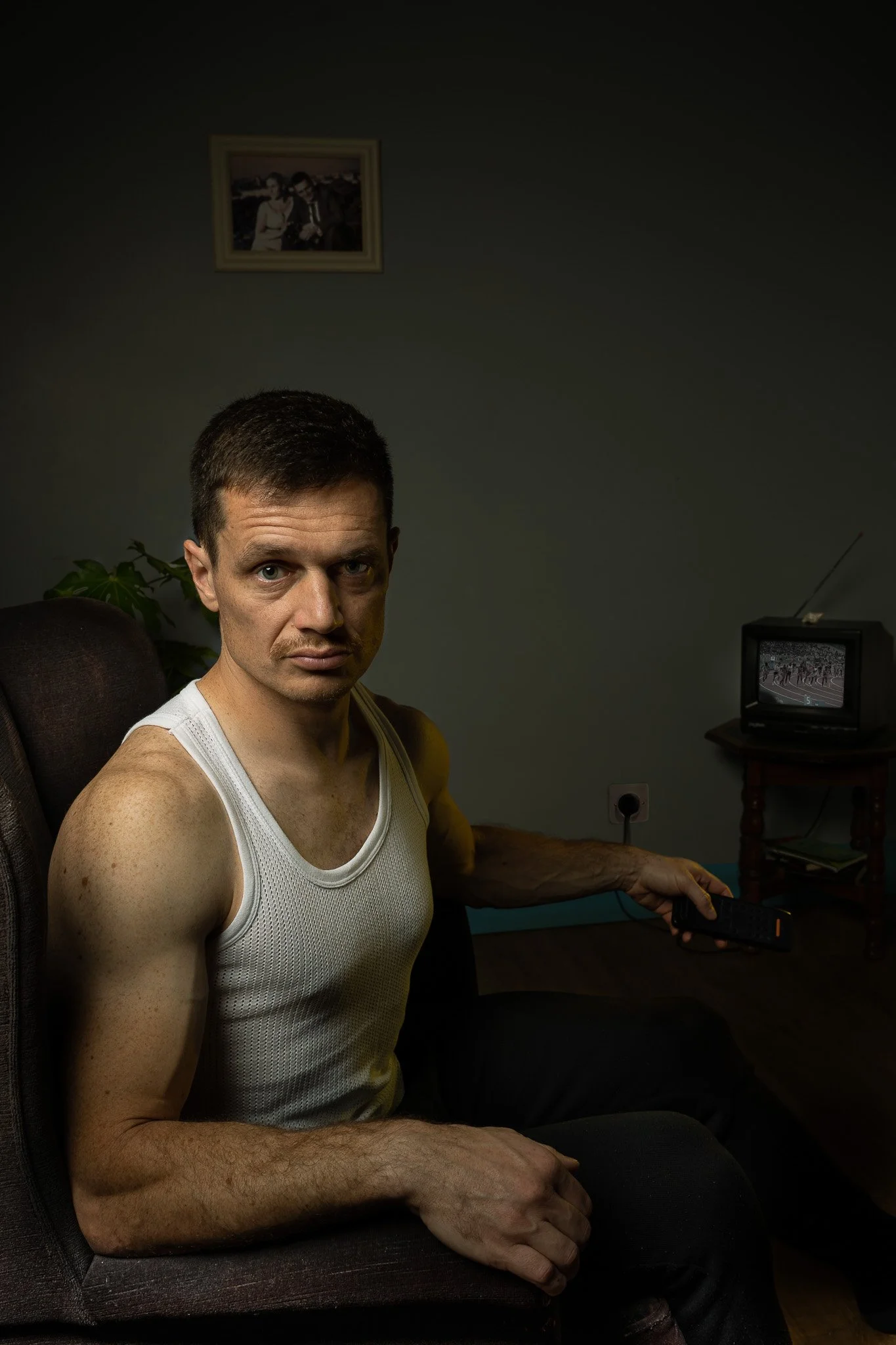 A man sitting on a dark sofa in a dimly lit room, holding a remote control, with an old television showing a race or sports event. A framed photo is hanging on the wall behind him.