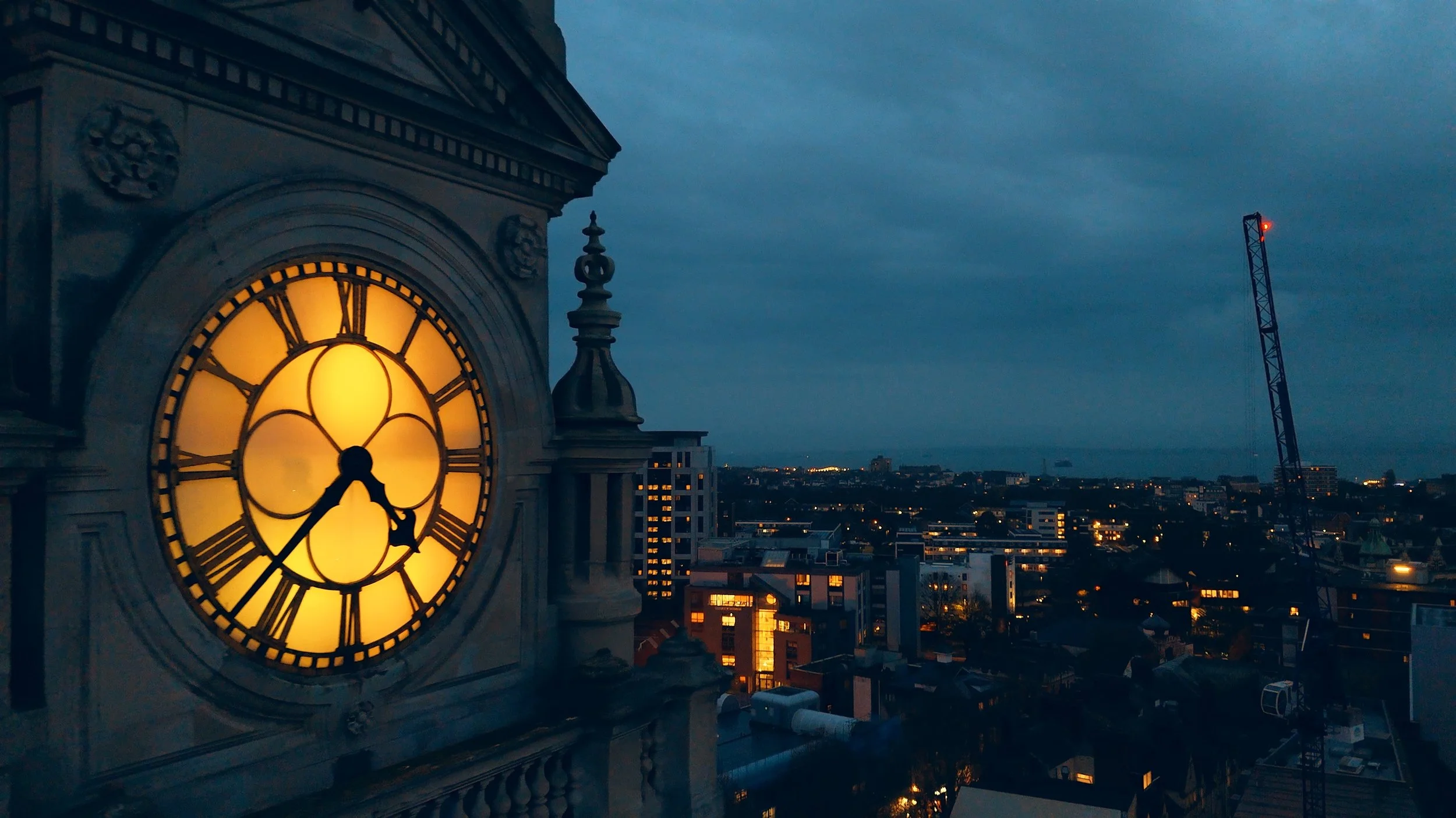 A large illuminated clock on a building at dusk shows the time as 8:29. The city skyline with buildings, streetlights, and a crane is visible in the background under a cloudy sky.