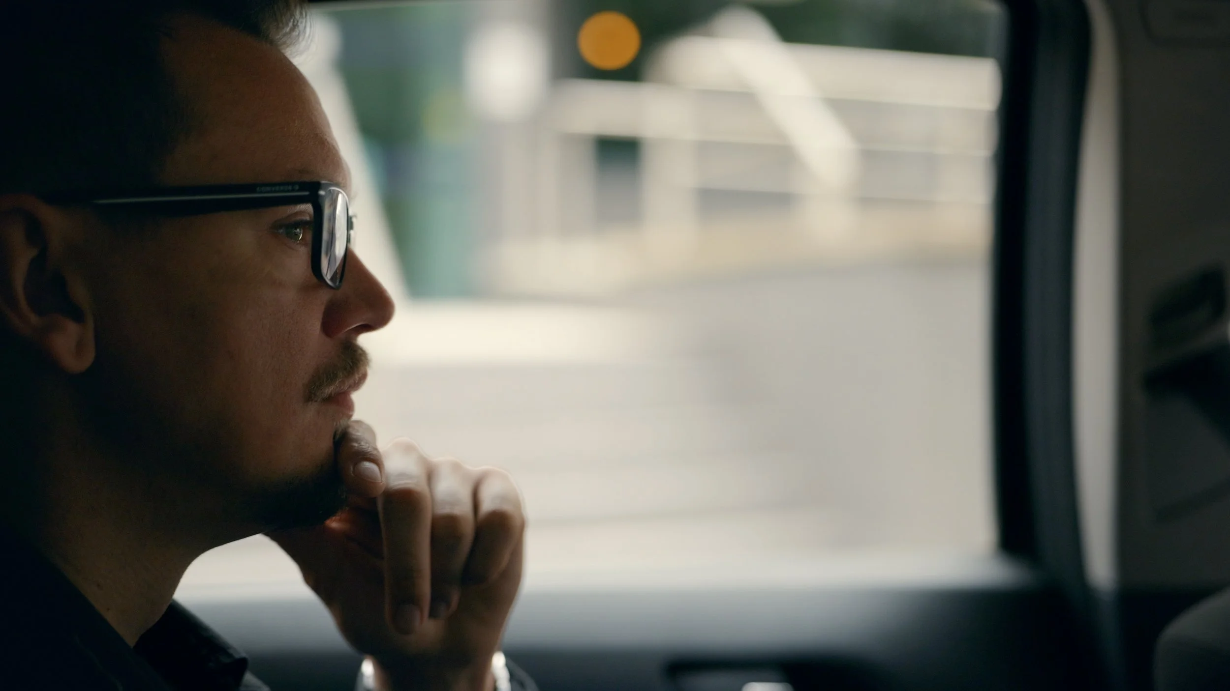 A man with glasses and a mustache sitting inside a vehicle, gazing thoughtfully with his chin resting on his hand.
