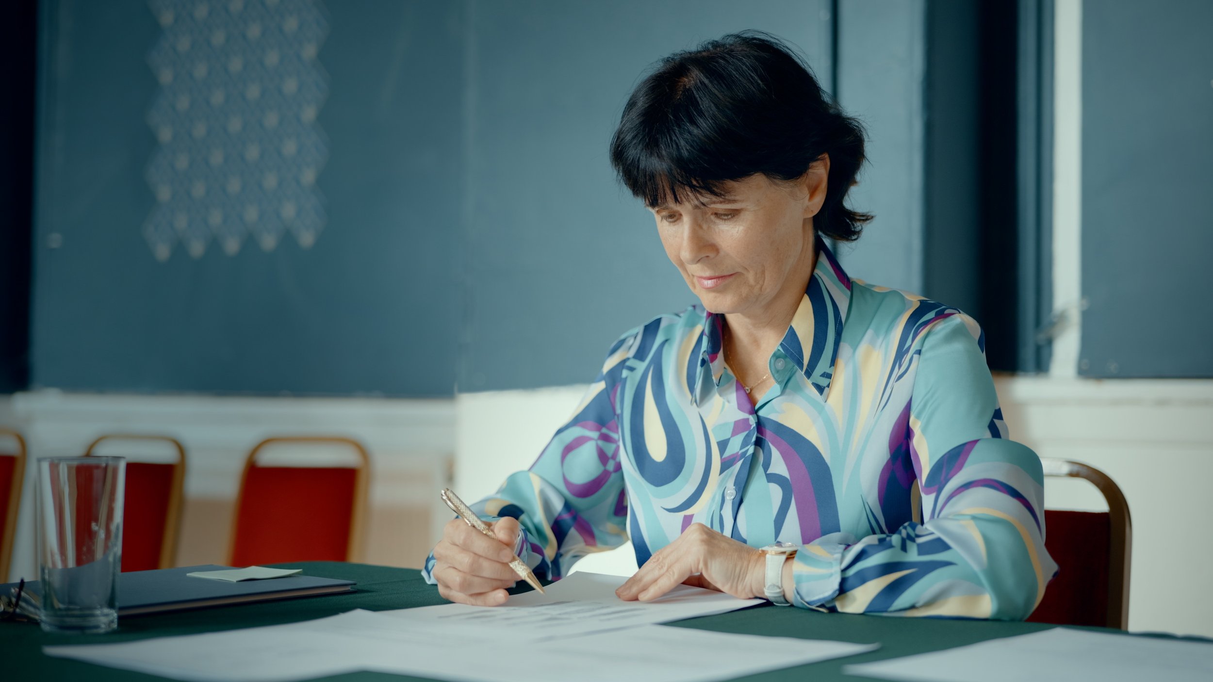 A woman with short black hair wearing a colorful patterned blouse sitting at a table, writing on papers with a pen, in a room with blue walls and windows.