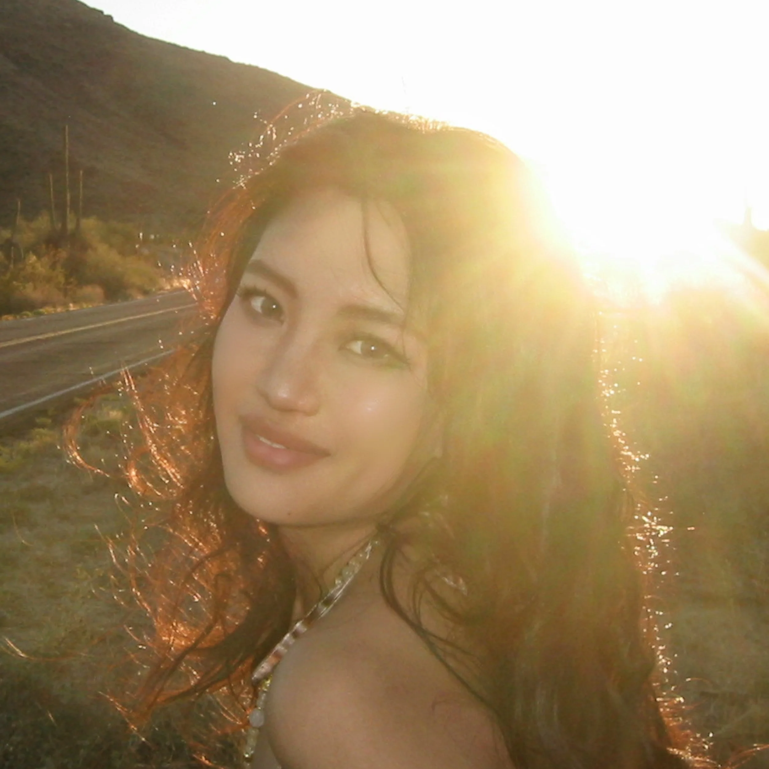 A woman with long, wavy hair smiling at the camera with the sunset behind her in a desert landscape with cacti and hills.
