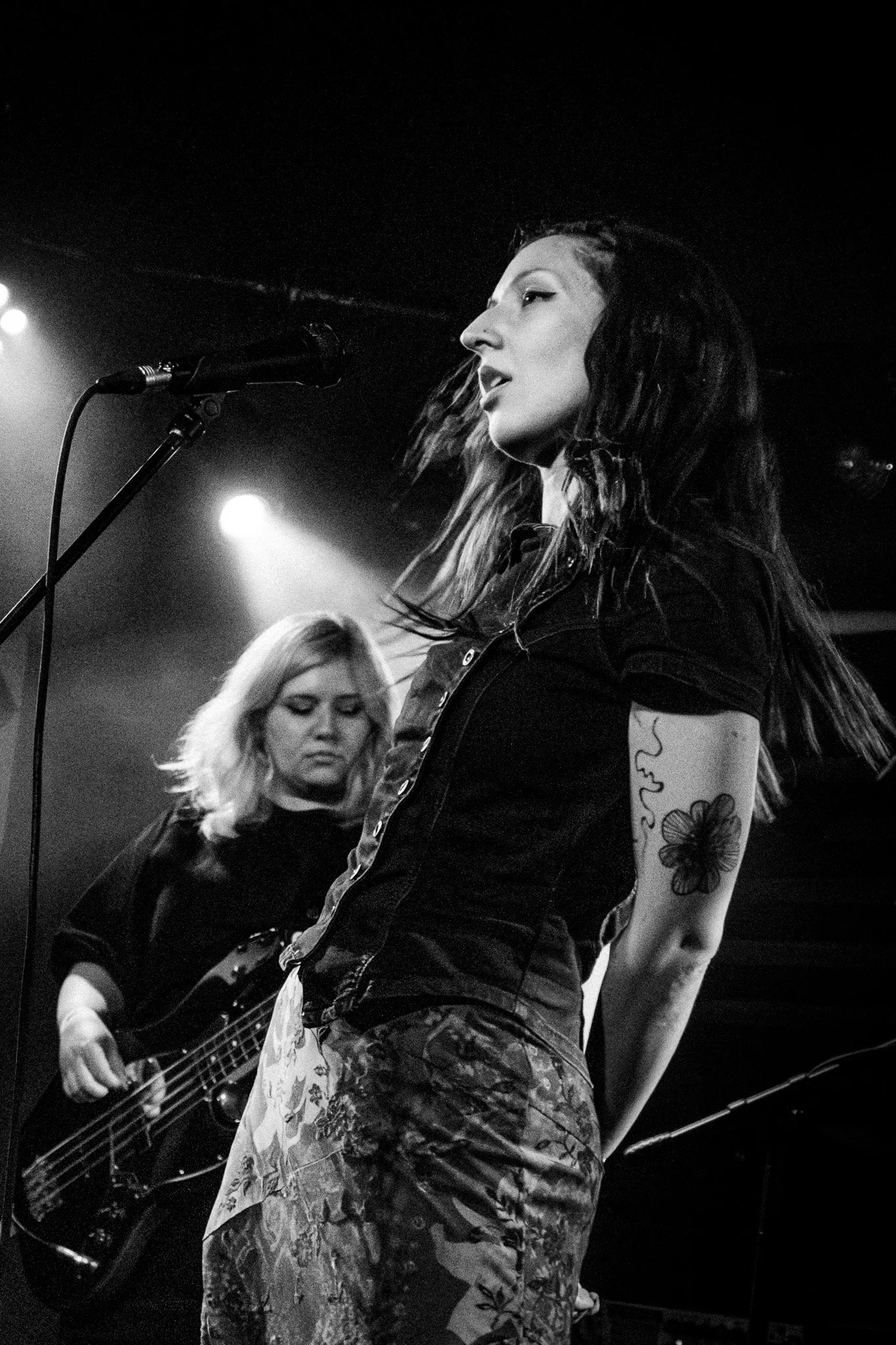 Black and white photo of a Maia singing into a microphone, and Paula playing bass in the background.