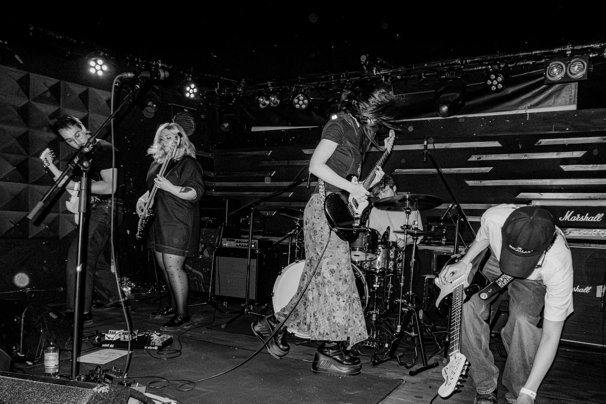 A live performance on stage featuring Maia and the Squires. The scene is in black and white with stage lights overhead and equipment on stage.