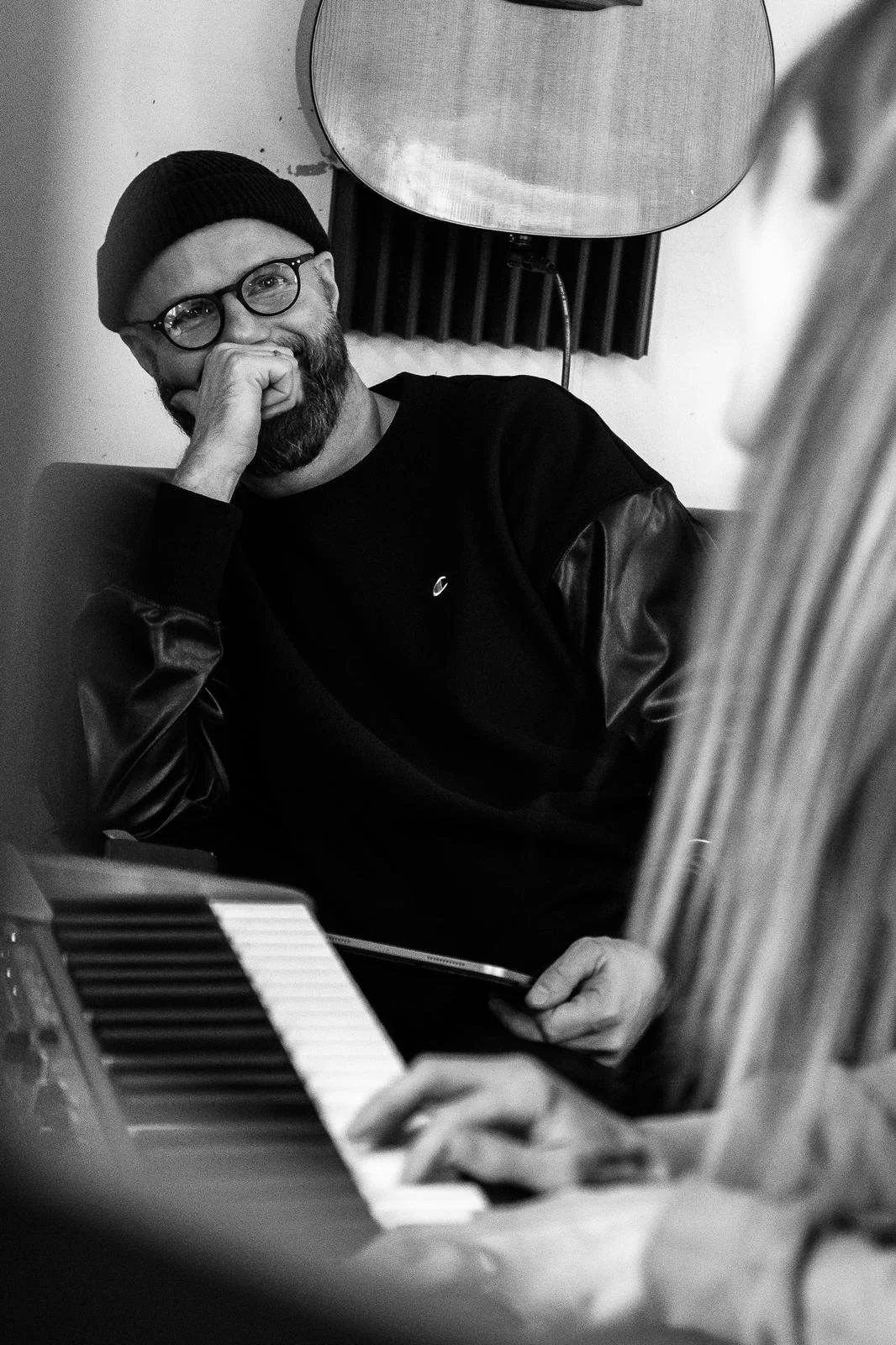 Man with glasses and beanie, smiling, seated near a keyboard, black and white photo.