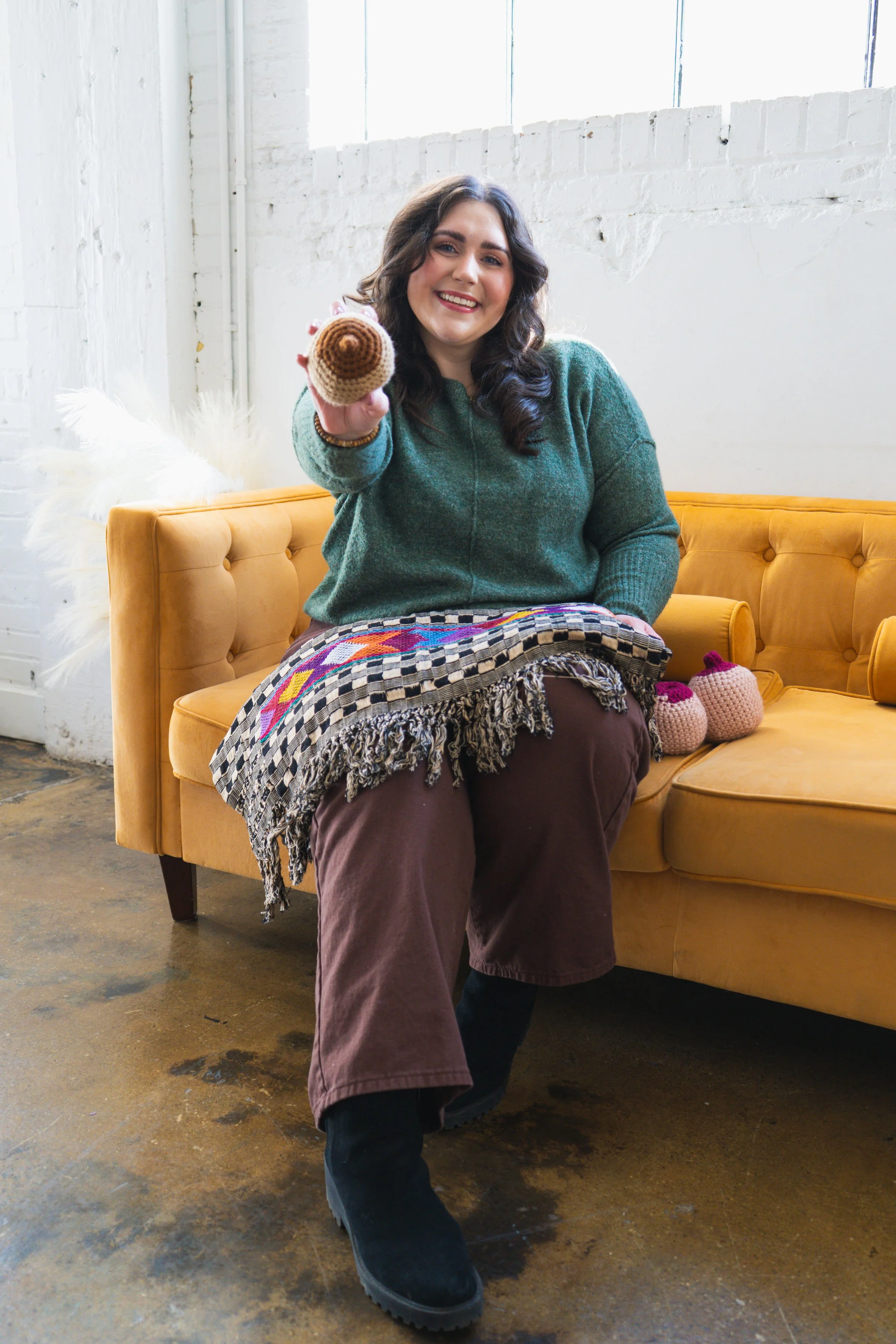 Doula sitting on yellow tufted couch holding up a crochet breast model toward the camera, smiling, with a colorful fringed blanket and additional breast models beside her