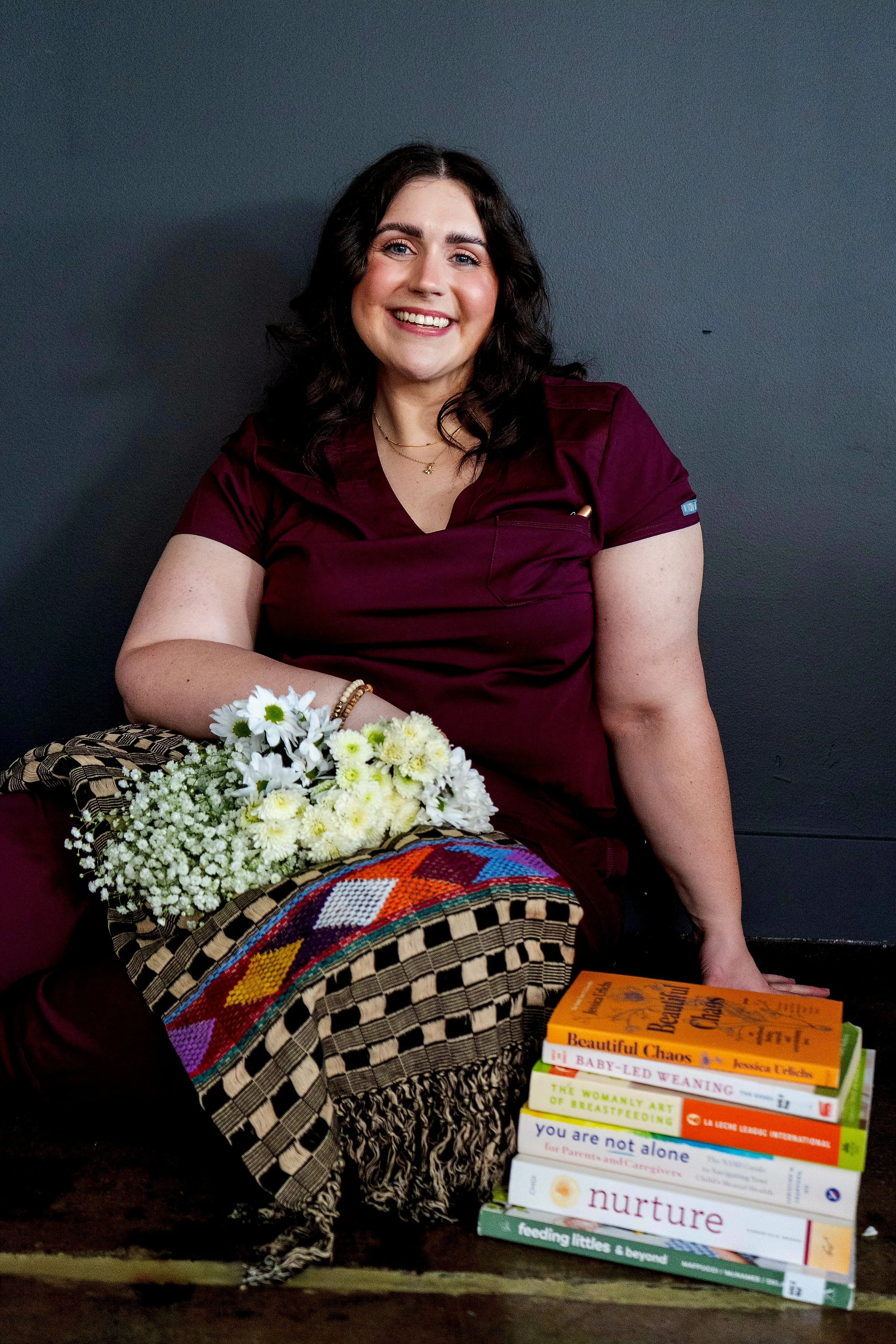 Jorie-Brae Morgan birth and postpartum doula smiling in burgundy scrubs holding white flowers beside stack of pregnancy and breastfeeding books including Beautiful Chaos, The Womanly Art of Breastfeeding, and Nurture