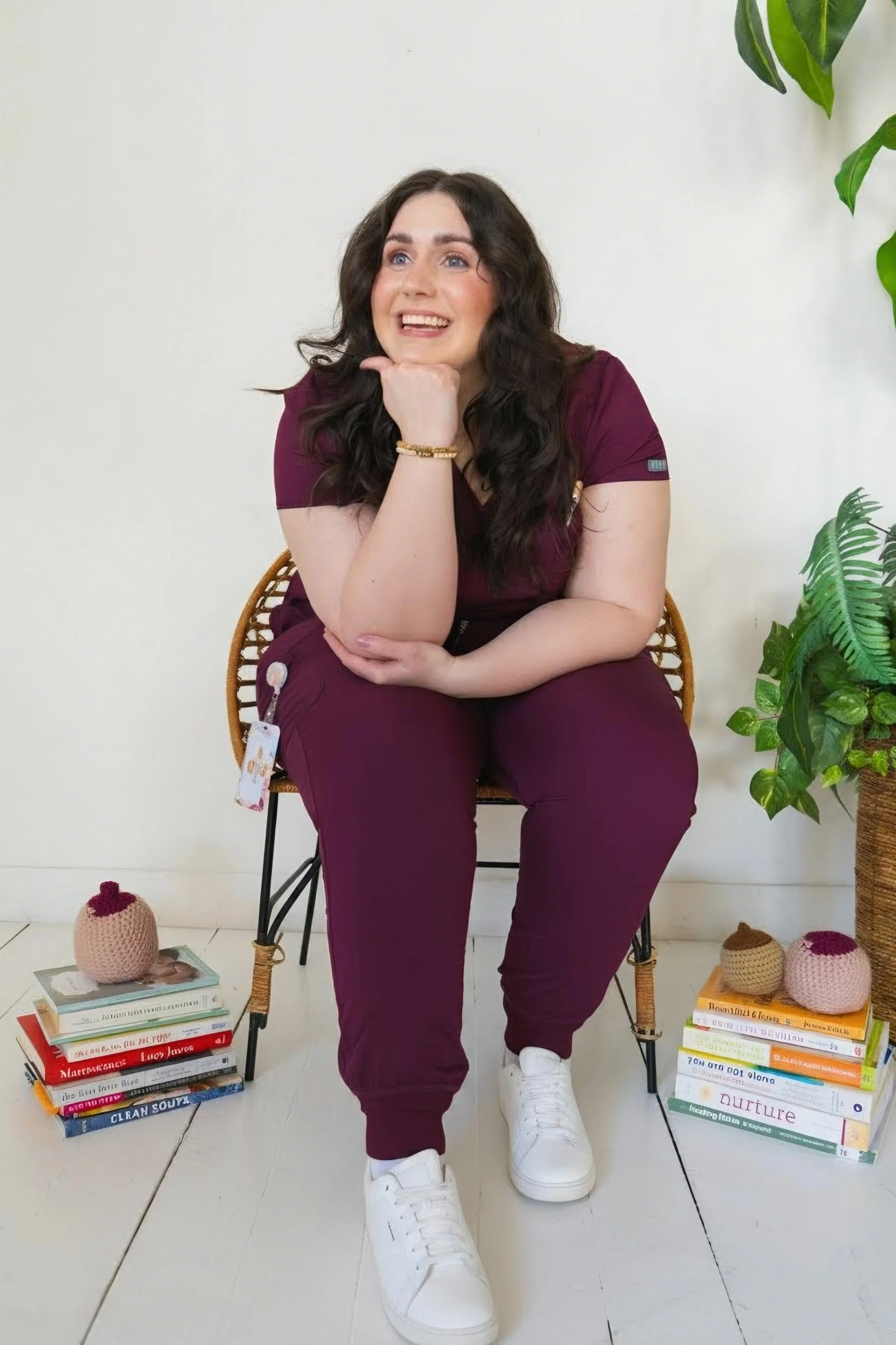 Doula in burgundy scrubs sitting in rattan chair, smiling and surrounded by stacks of birth and breastfeeding books with crochet breast models