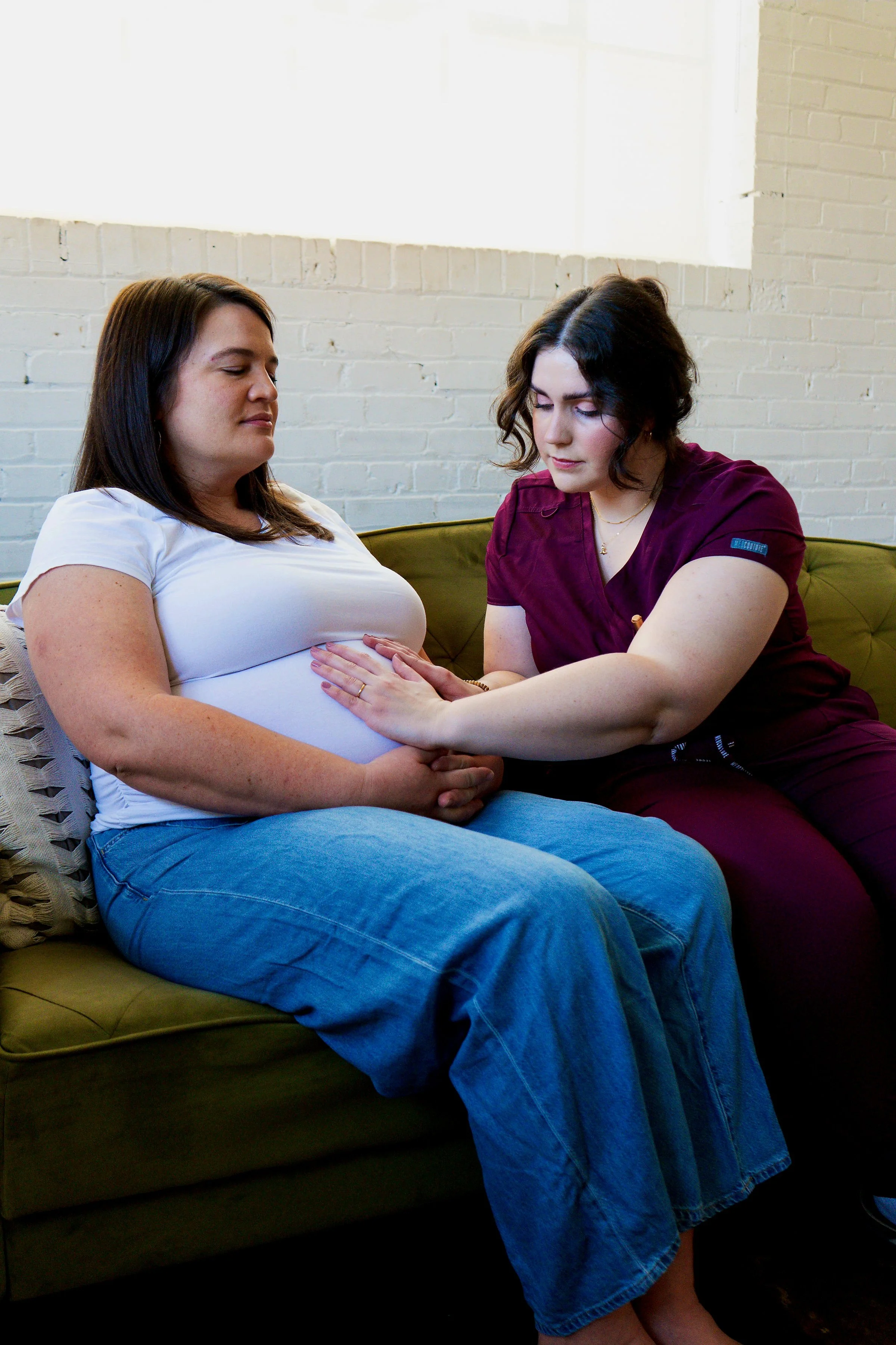 Doula sitting beside pregnant client on olive couch, gently placing hand on baby bump during prenatal consultation