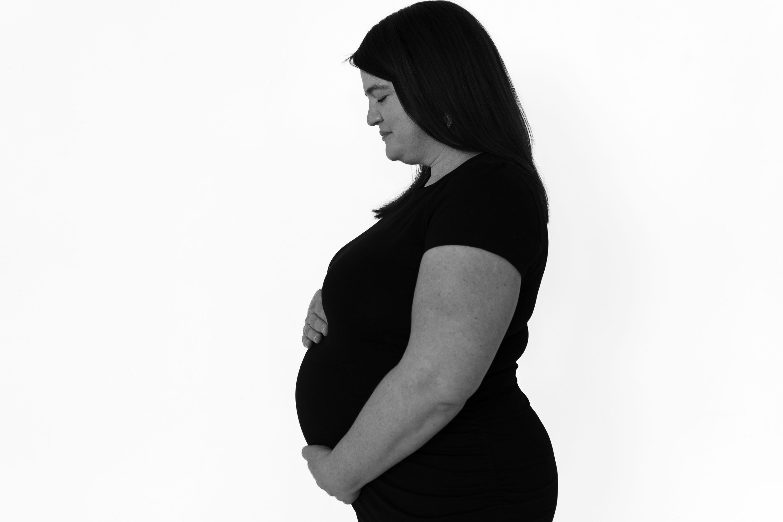 Black and white side profile of pregnant woman in black dress cradling her baby bump, looking down with a calm expression