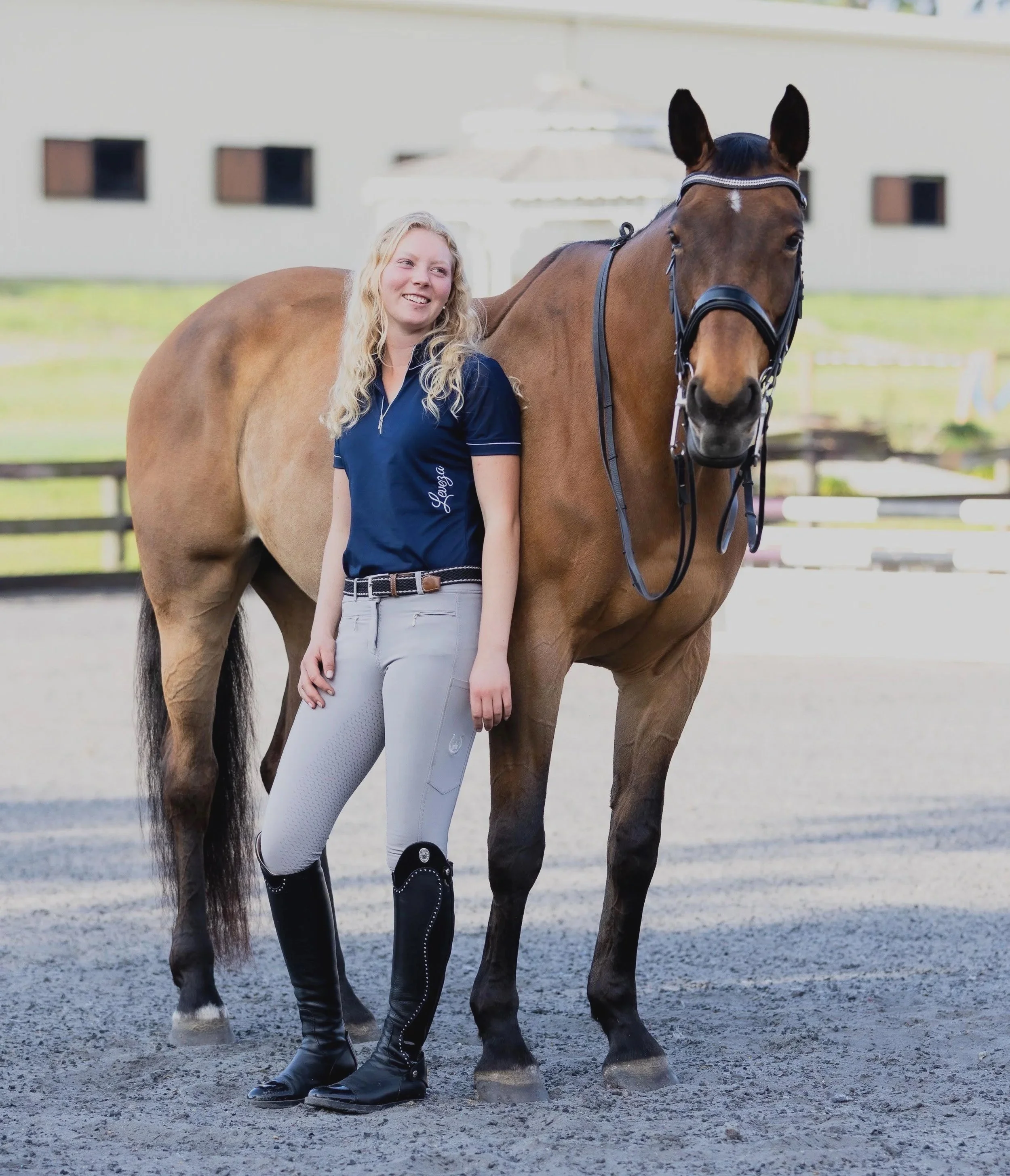 A woman with long blonde hair wearing a navy polo shirt, white riding pants, and black riding boots standing next to a brown horse with a black mane outside on a riding arena.
