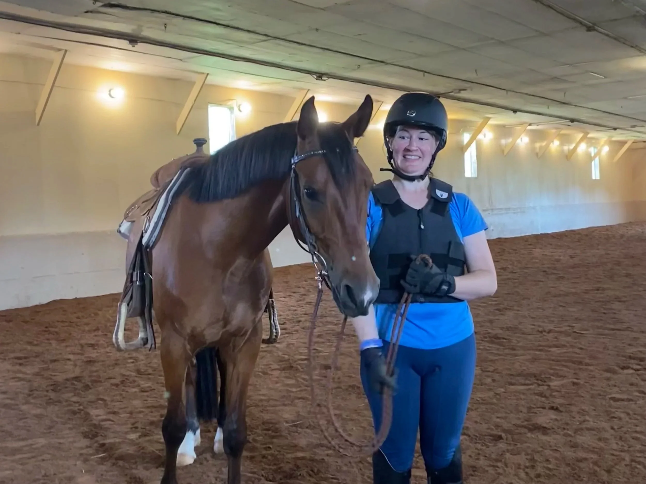 A smiling woman wearing a black riding helmet and blue shirt stands beside a brown horse inside an indoor riding arena. The woman holds the horse's reins, and the horse has a saddle on its back.
