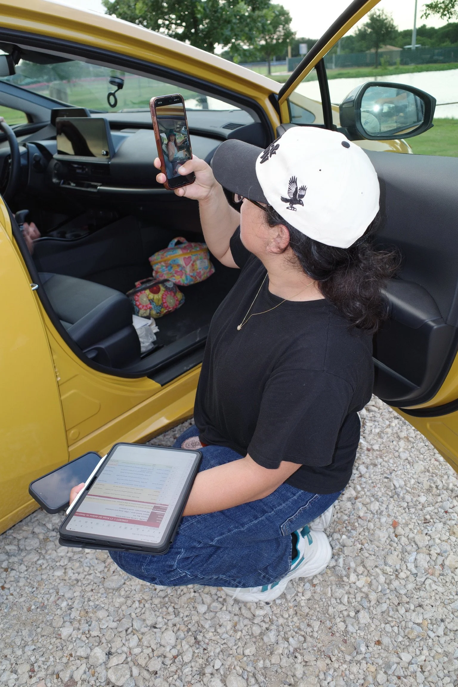 A woman wearing a white hat with black bird illustrations is kneeling on gravel ground outside next to a yellow car. She is holding a tablet with medication information and a phone taking a photo of a woman in the car. The car's interior includes a dashboard, steering wheel, and some colorful bags on the passenger seat. There is a body of water and trees in the background.