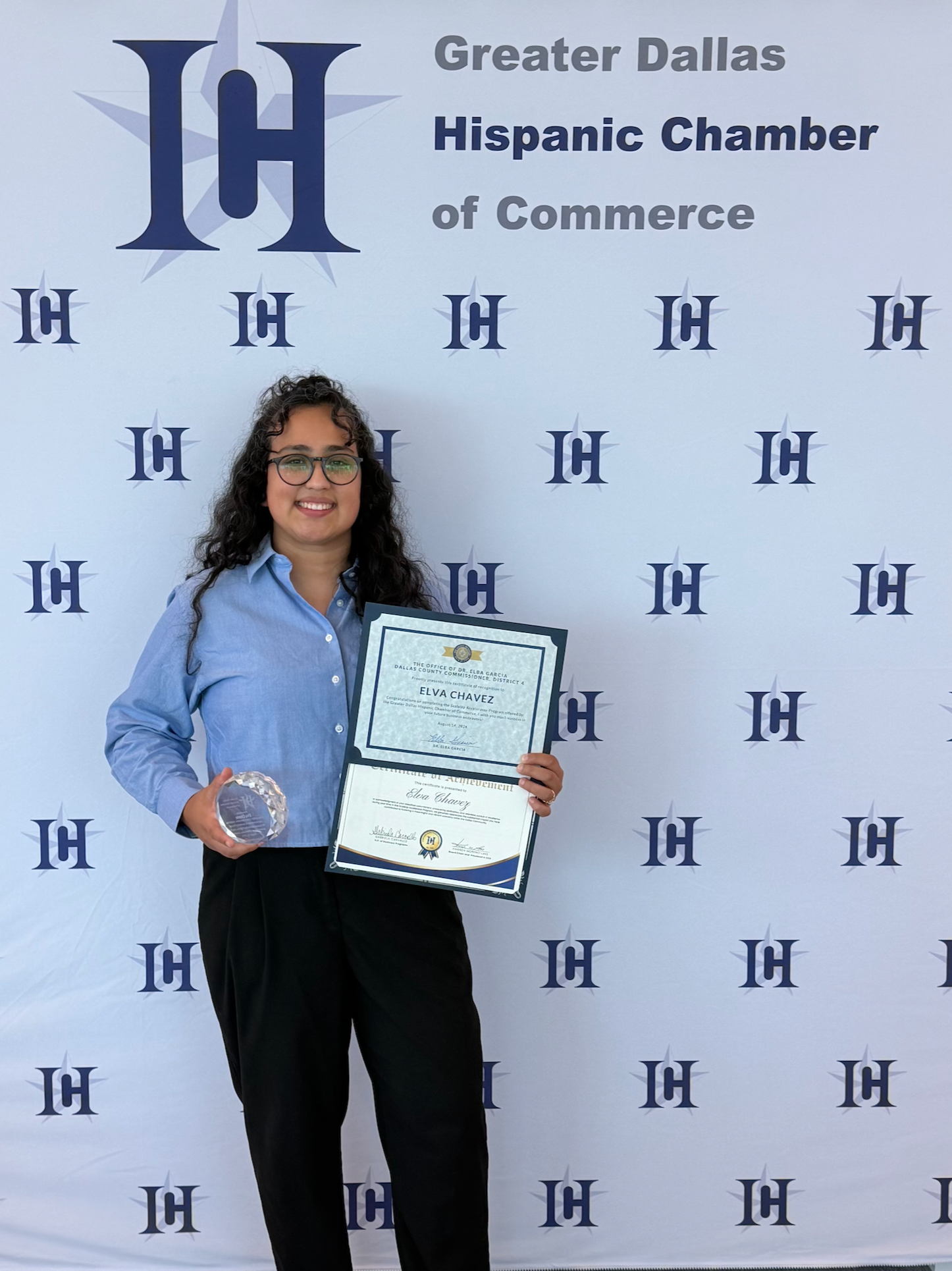 A woman standing in front of a backdrop with the logo of Greater Dallas Hispanic Chamber of Commerce. She is smiling, wearing glasses, a blue shirt, and black pants. She is holding a certificate and a glass award.