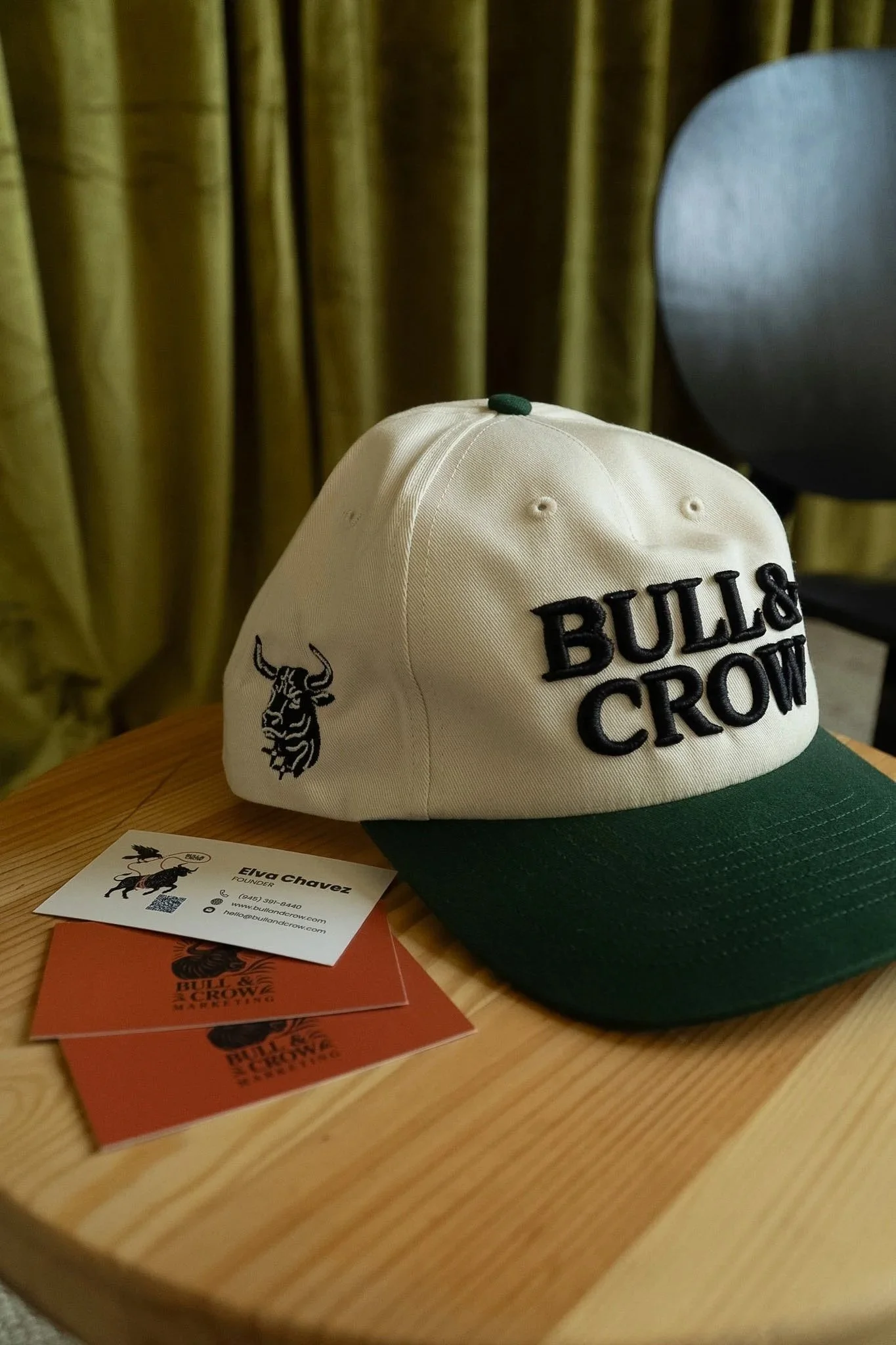 A white and green baseball cap with the words 'BULL & CROW' embroidered on the front, placed on a wooden table next to business cards and orange pamphlets on a wooden table, with a black chair and a yellow curtain in the background.