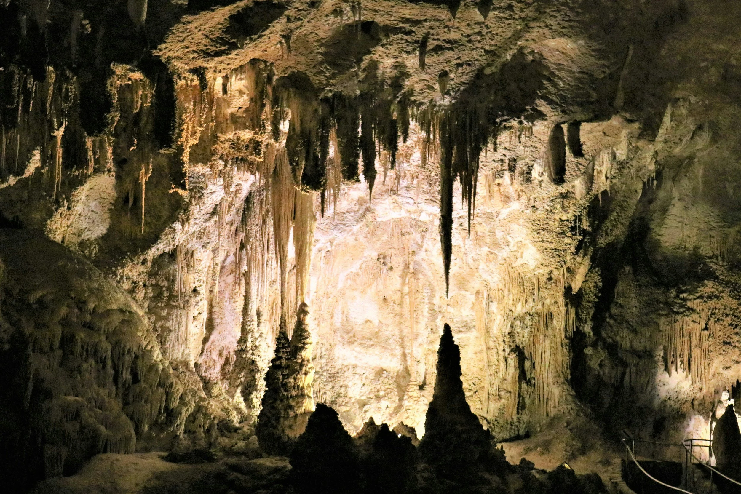 Cave interior with stalactites and stalagmites, featuring illuminated rock formations and a railing path.