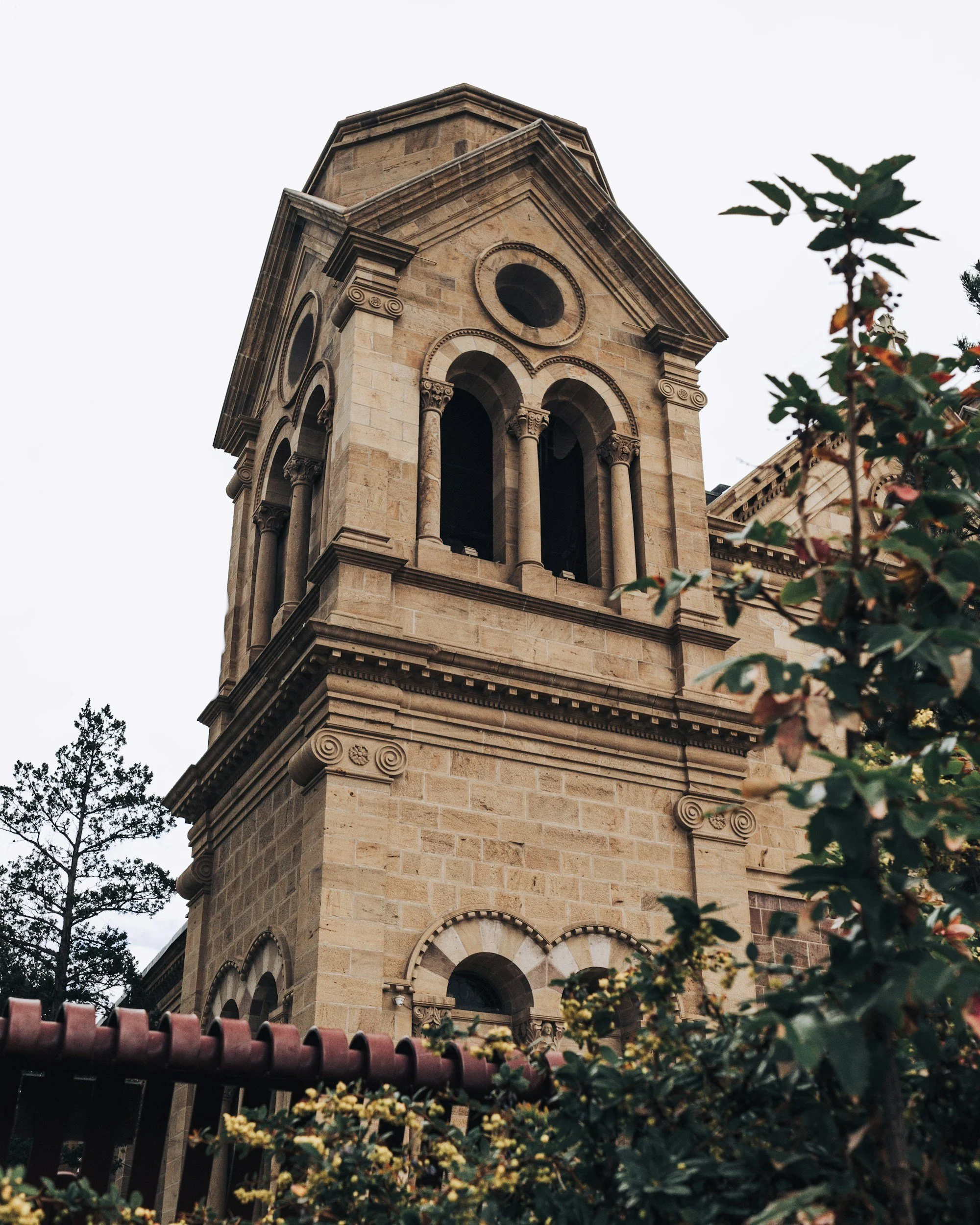 Historic stone church tower with arched windows and decorative architectural details, surrounded by greenery and a cloudy sky in the background.