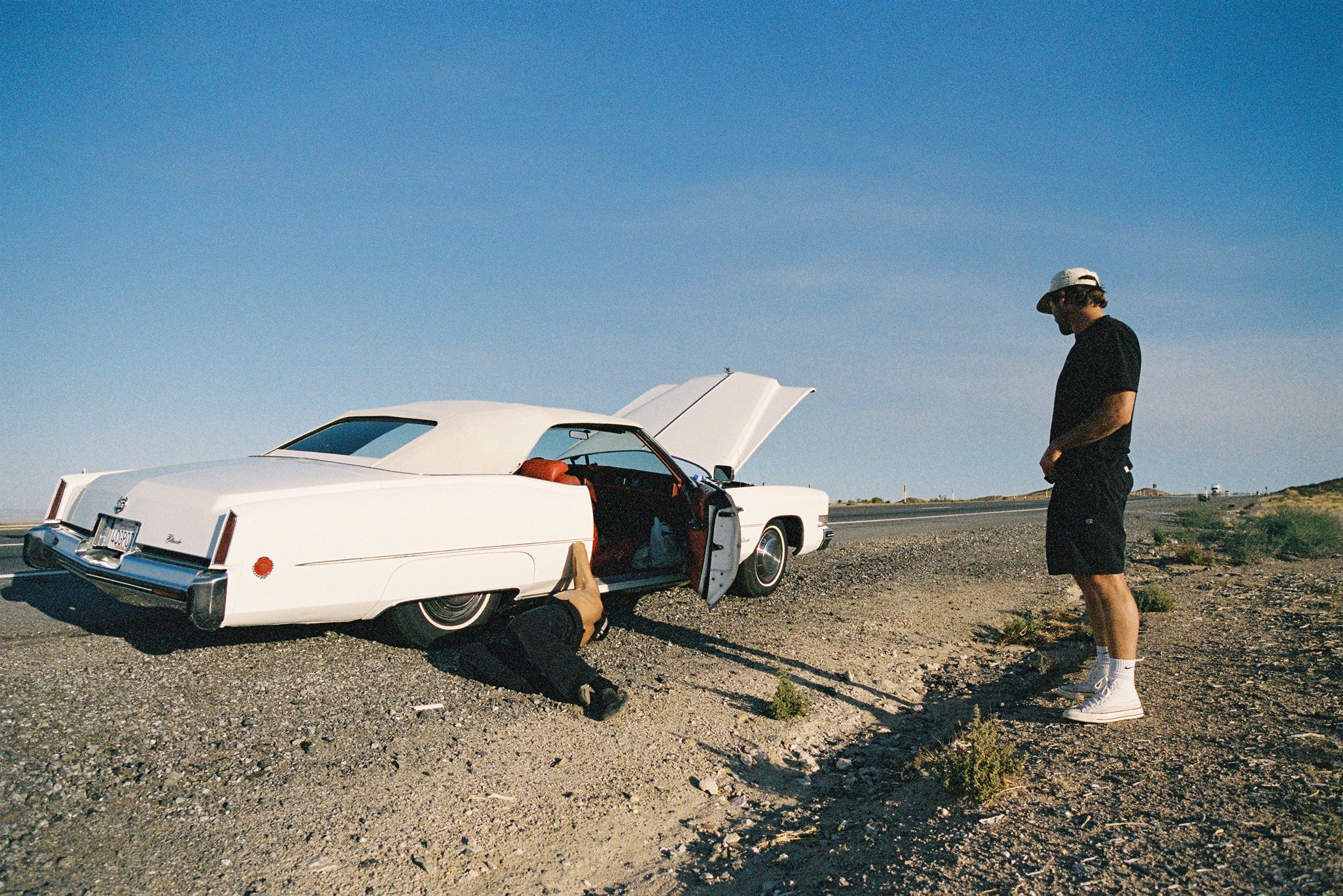 A person lying under a white classic car with an open hood, and another person standing nearby on the side of a desert road.