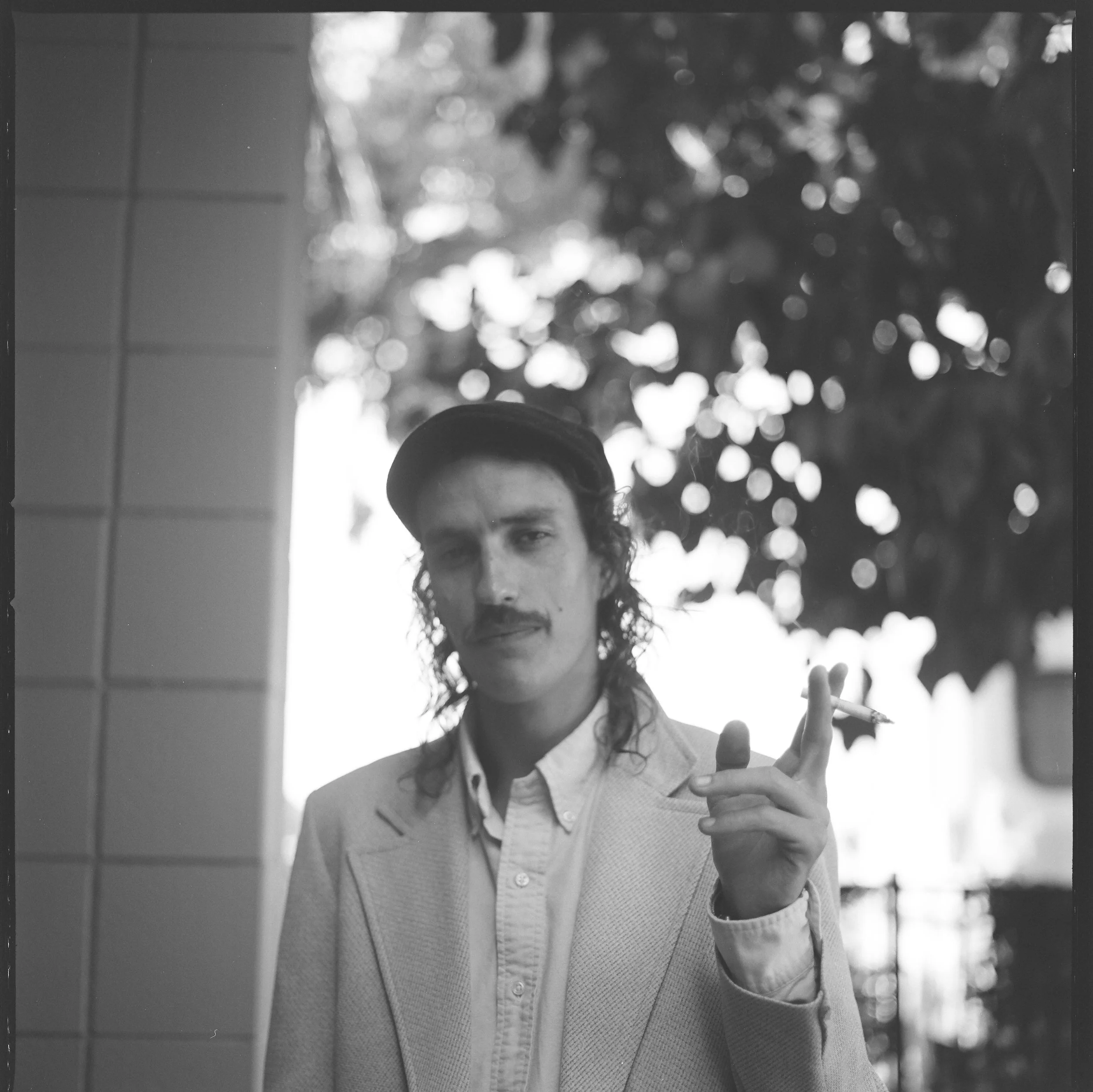 Black and white portrait of a person with long hair and a mustache, wearing a cap, light-colored jacket, and holding a cigarette, standing outdoors with trees in the background.