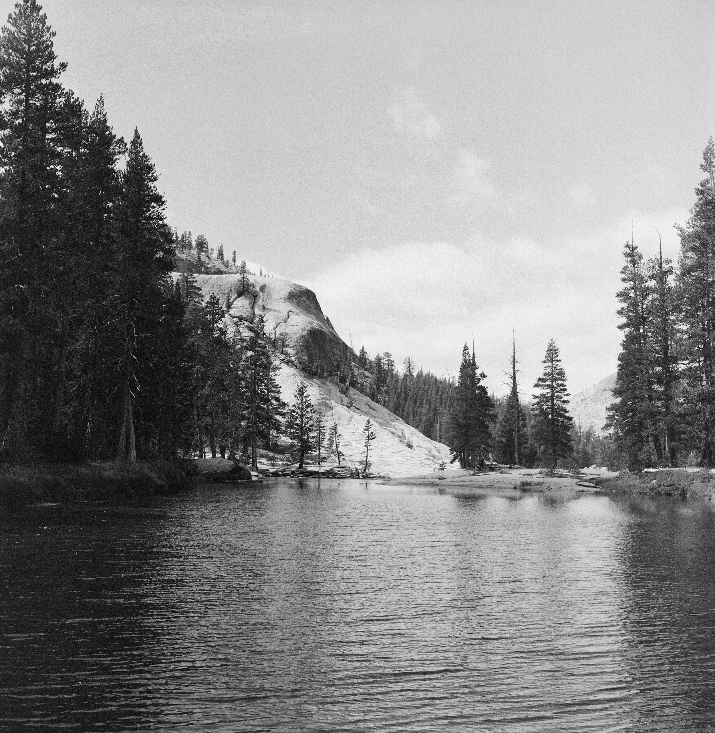 Black and white photo of a serene lake surrounded by pine trees and rocky cliffs under a cloudy sky.