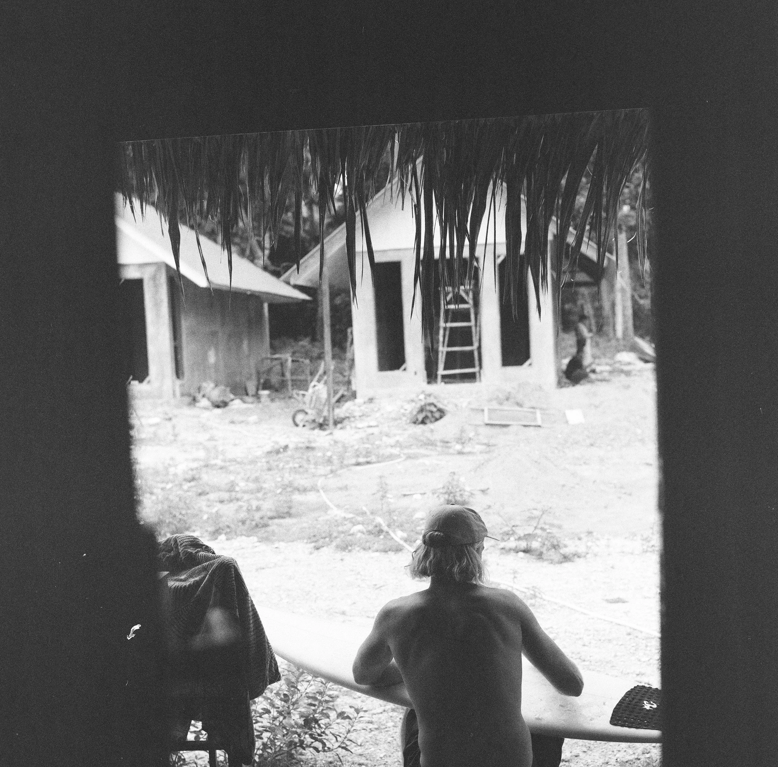 A person sitting near a surfboard, with two unfinished huts in the background, surrounded by a construction site. The scene is monochrome.