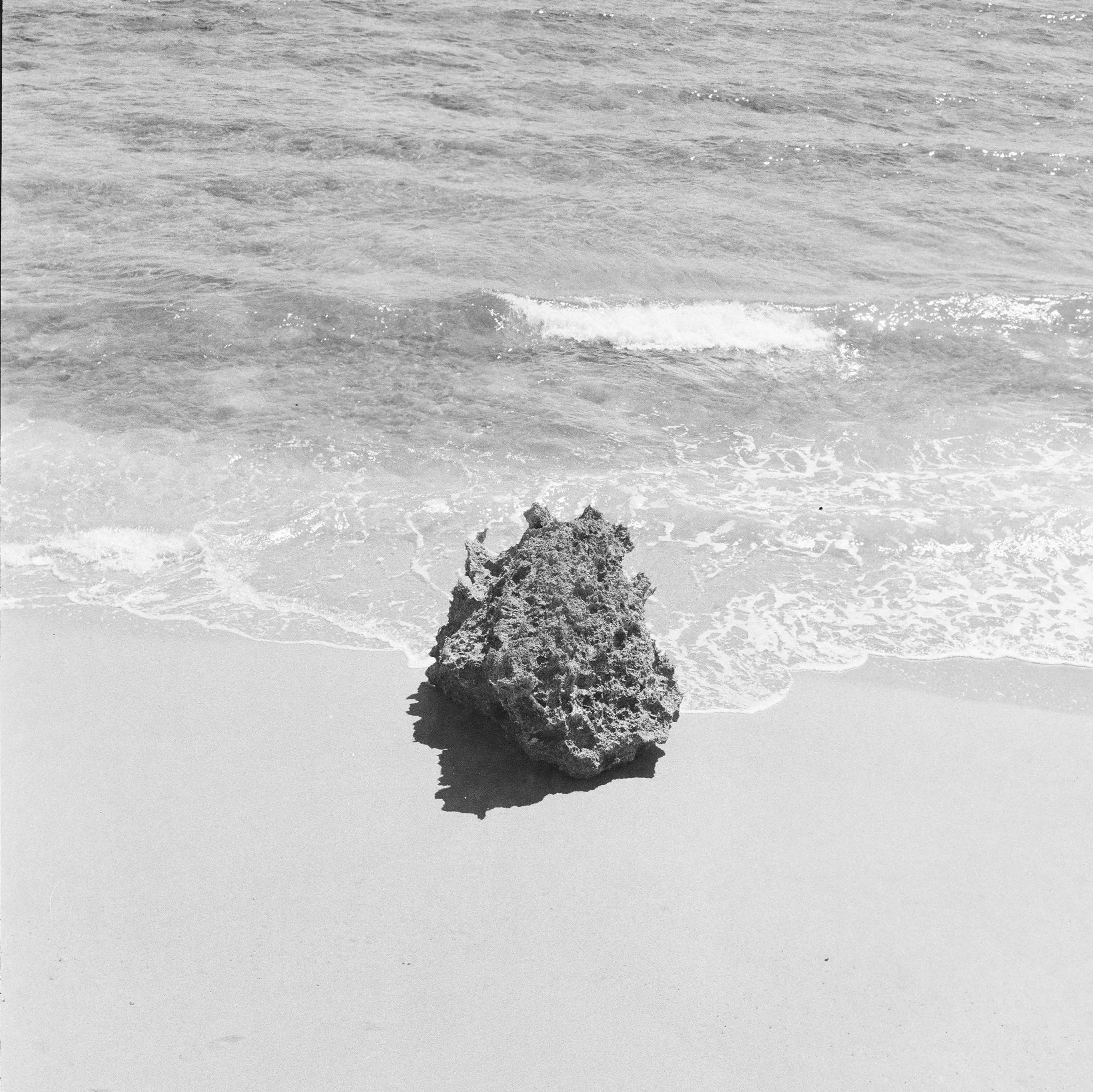 Black and white photo of a rocky beach with a large rock on the sand near the shoreline.