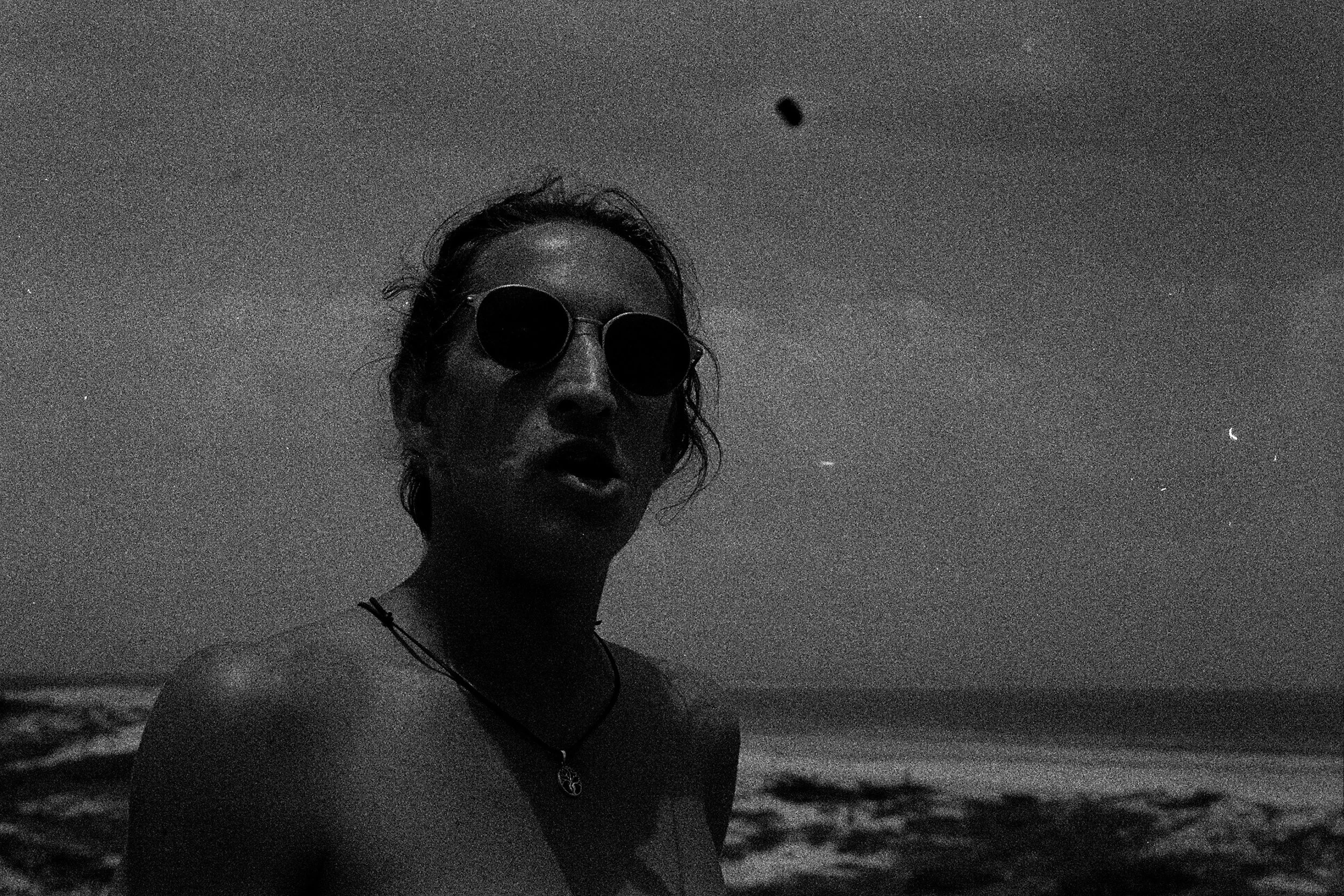 Black and white photo of a person wearing sunglasses at the beach, taken at an upward angle showing the sky.