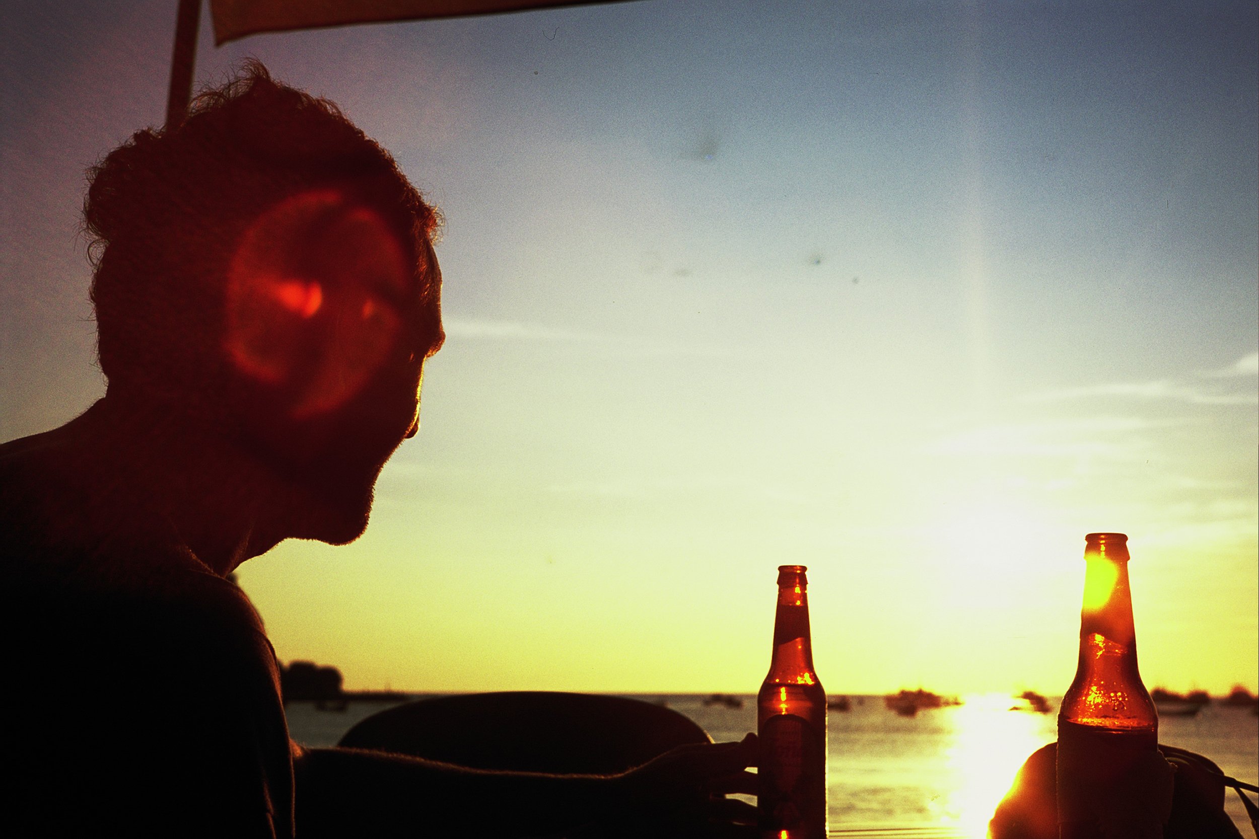 Silhouette of a person holding a bottle with a sunset in the background, two bottles on a table, and a glowing sky over a body of water.