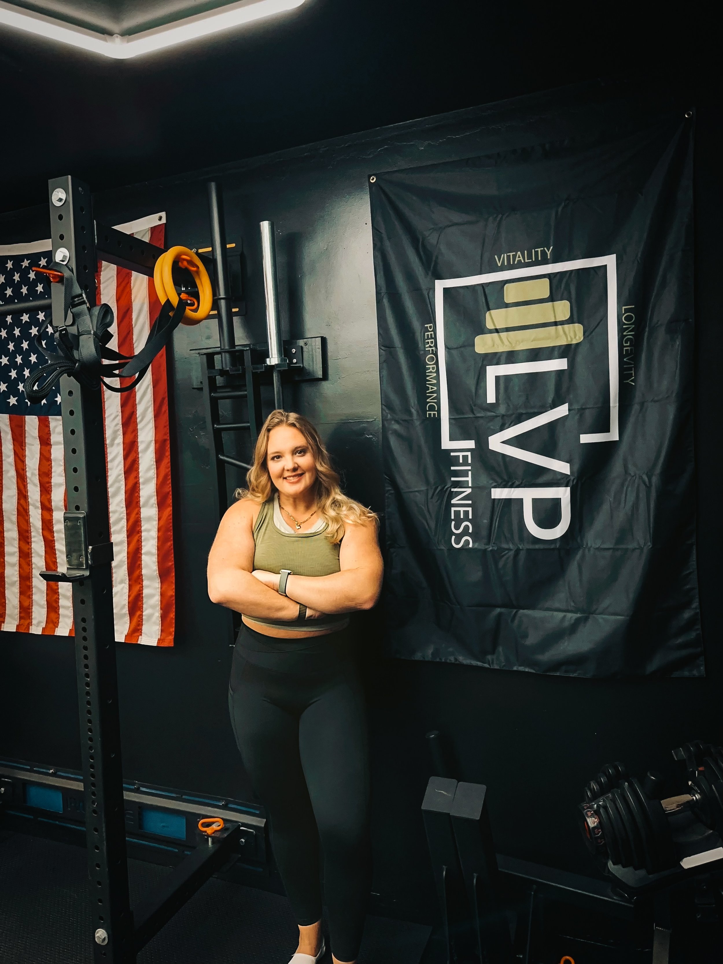 A woman standing in a gym with her arms crossed, smiling, next to a black banner with the logo 'LVP Fitness' and an American flag in the background.