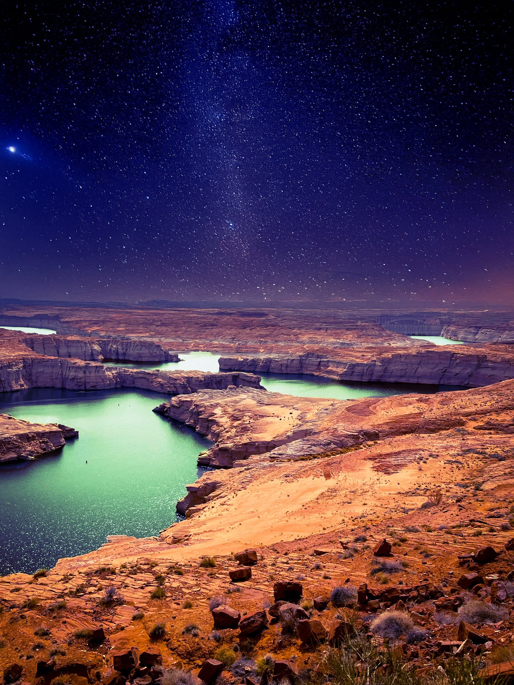 Starry night sky over a desert landscape with a river or lake running through reddish canyon cliffs.