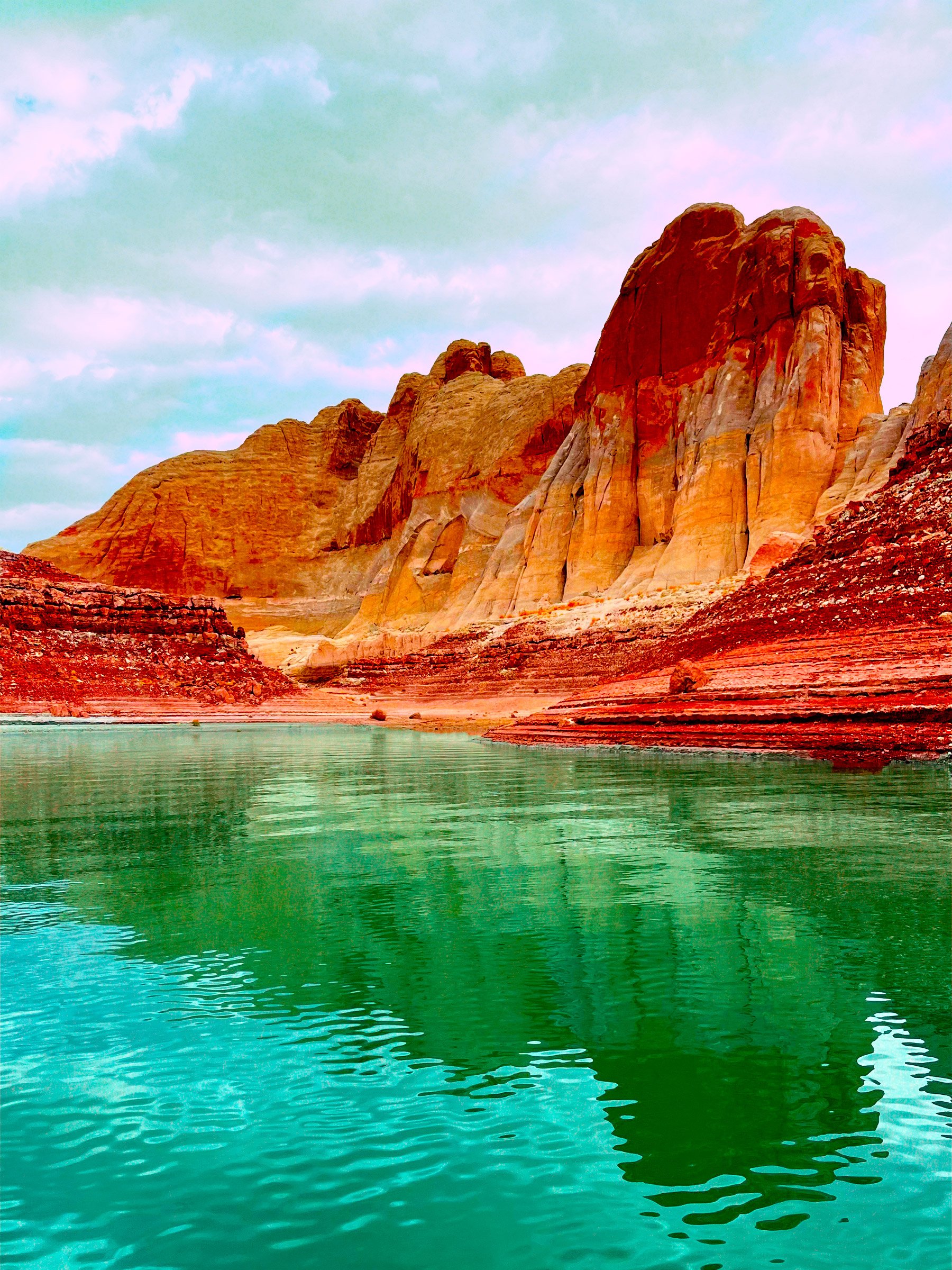 Colorful desert landscape with red-orange rock formations and green water in the foreground under a partly cloudy sky.