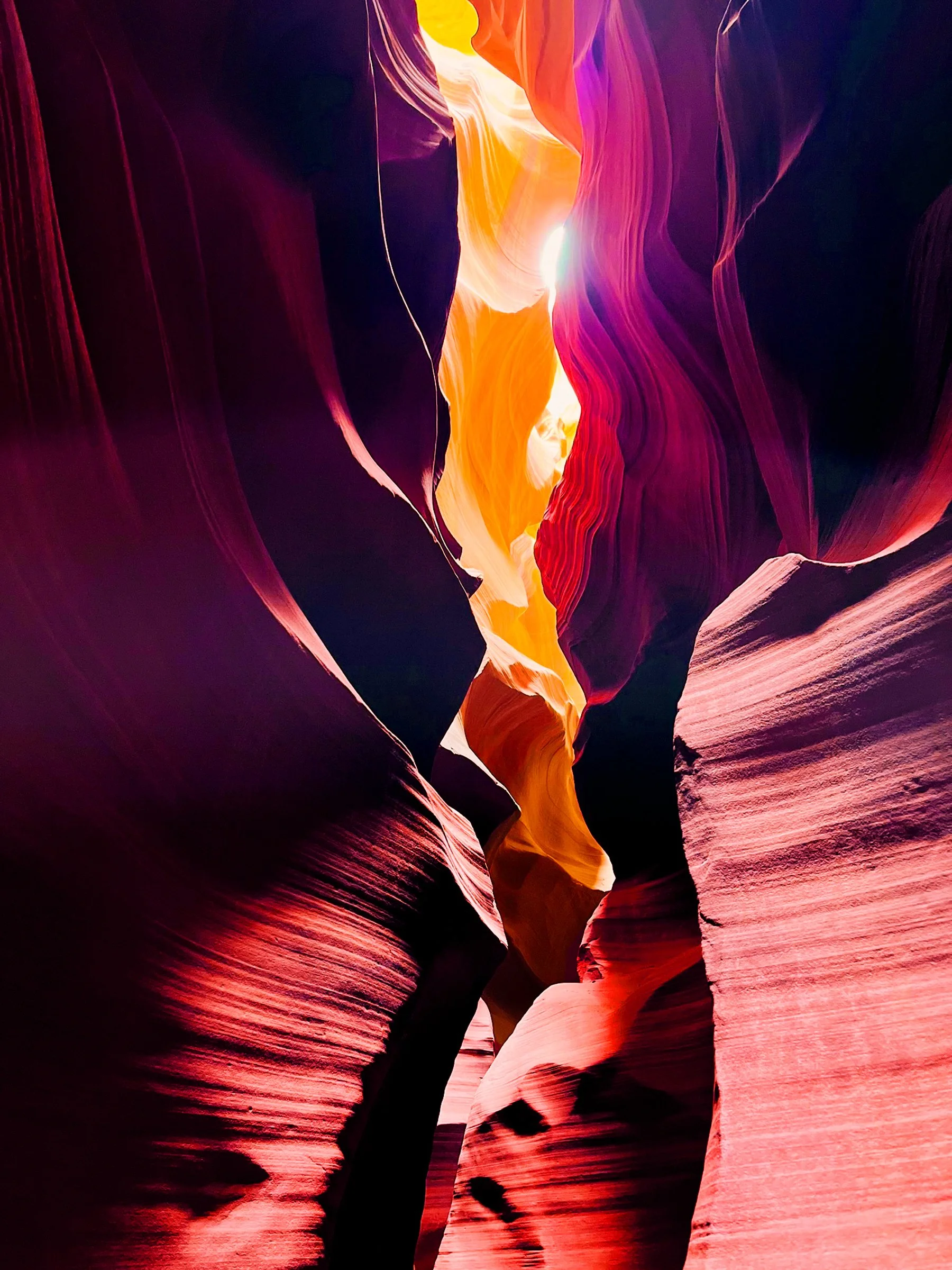 Colorful slot canyon with smooth, flowing rock formations in shades of purple, red, orange, and yellow, with sunlight filtering through at the top.