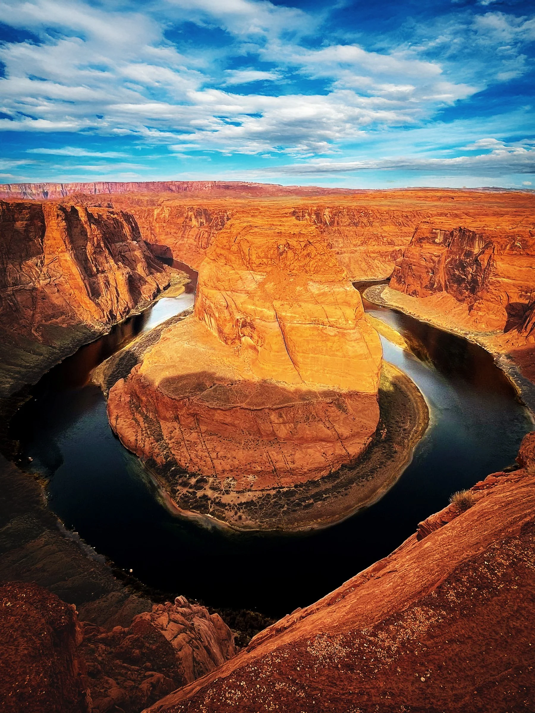 Aerial view of the Horseshoe Bend in the Colorado River, showing a U-shaped meander of the river surrounded by red rock cliffs under a partly cloudy blue sky.
