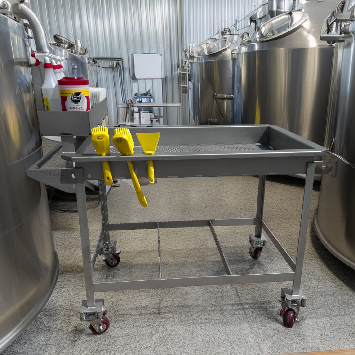 A mobile stainless steel cart is positioned in a clean industrial room with large metal tanks in the background, suggesting a food processing environment.