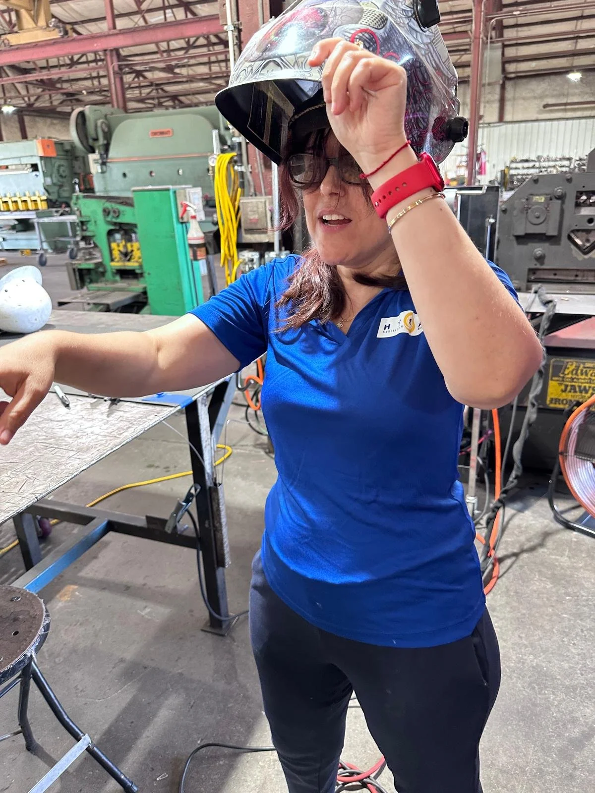 A woman stands at the center of a manufacturing workshop, wearing a blue shirt and black pants. She lifts her welding helmet with her left hand, revealing her face, and points toward something out of frame with her right hand.
